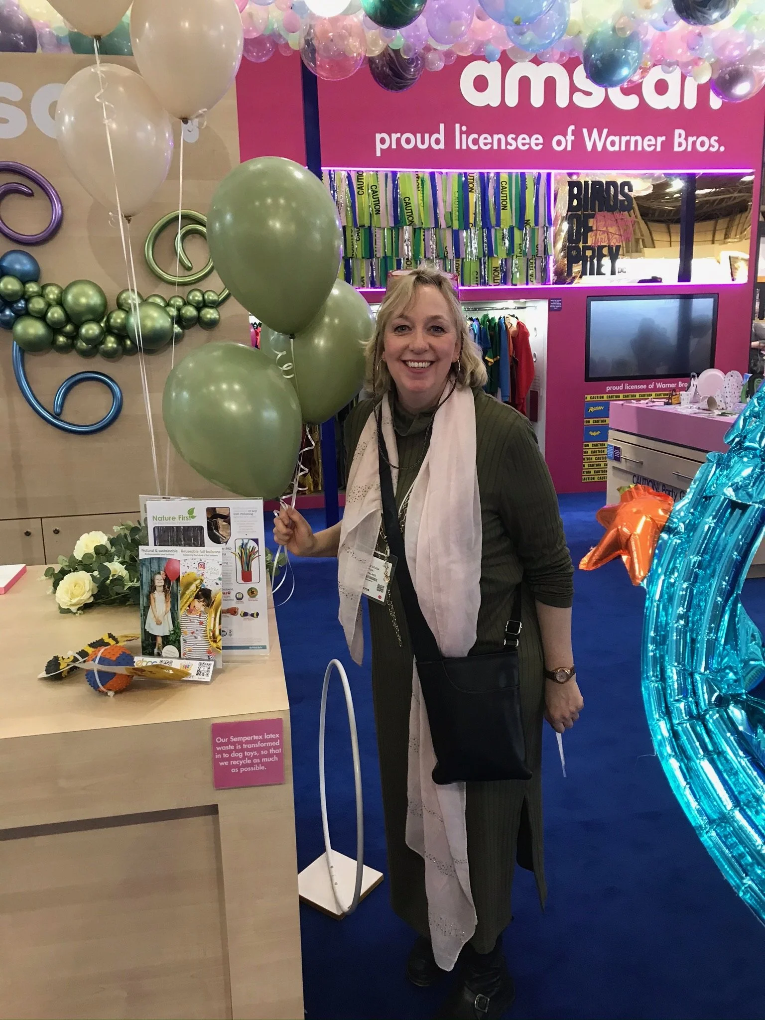 A woman holding gold and white balloons at an event booth with colorful decorations and a pink sign that says 'Aston' and 'proud licensee of Warner Bros.'