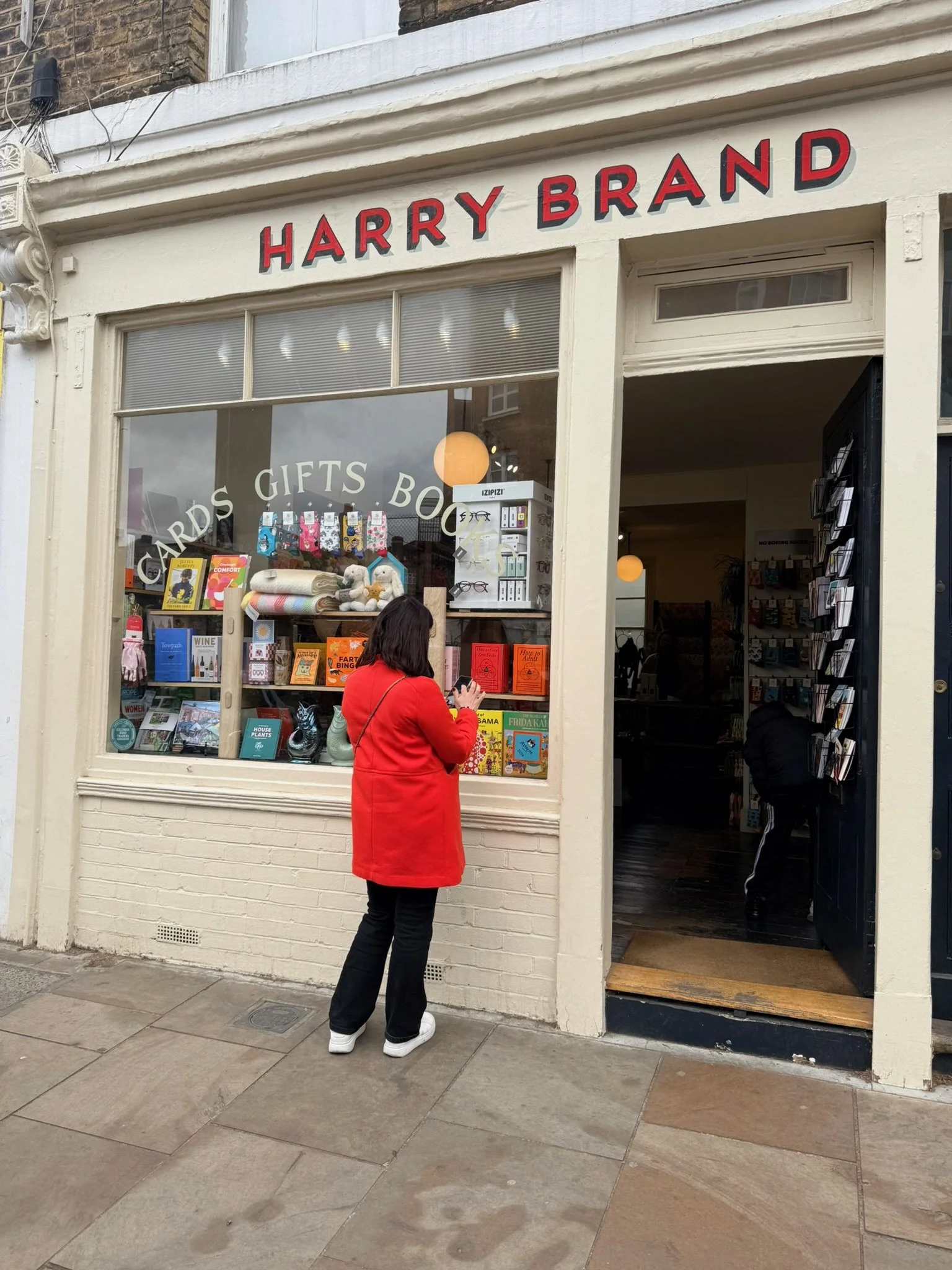 A person in a red coat stands outside a bookstore named Harry Brand, looking at the window display with various books and plush toys. The bookstore entrance is open, revealing more books inside.