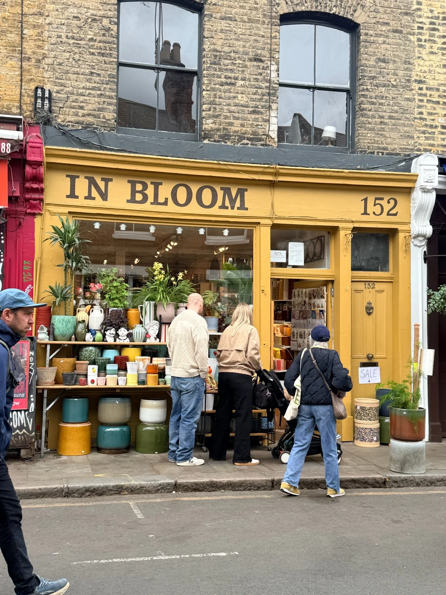 Street view of an outdoor shop with a yellow storefront labeled 'IN BLOOM' and the number '152'. The shop displays various pottery, planters, and decorative items. Several people are walking by or browsing in front of the shop, with some looking at t