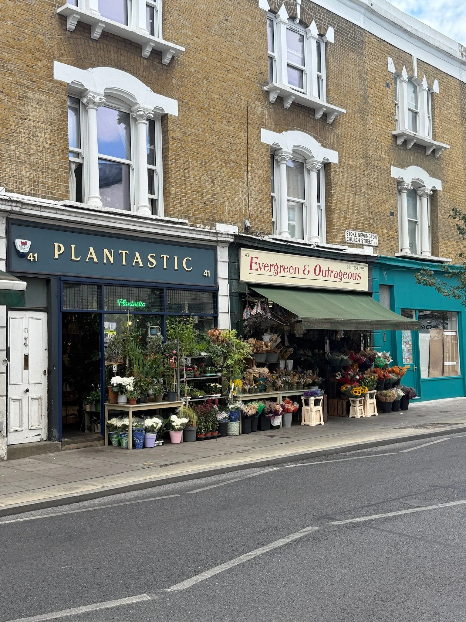 Street view of a flower shop with an array of potted flowers and plants outside. The shop has a dark blue facade with the name 'Plantastic' and a sign for 'Evergreen & Outrageous'. Above the shop, there are three-story brown brick buildings with white decorative window frames and trim. A street sign reads 'Stoke Newington Church Street'.