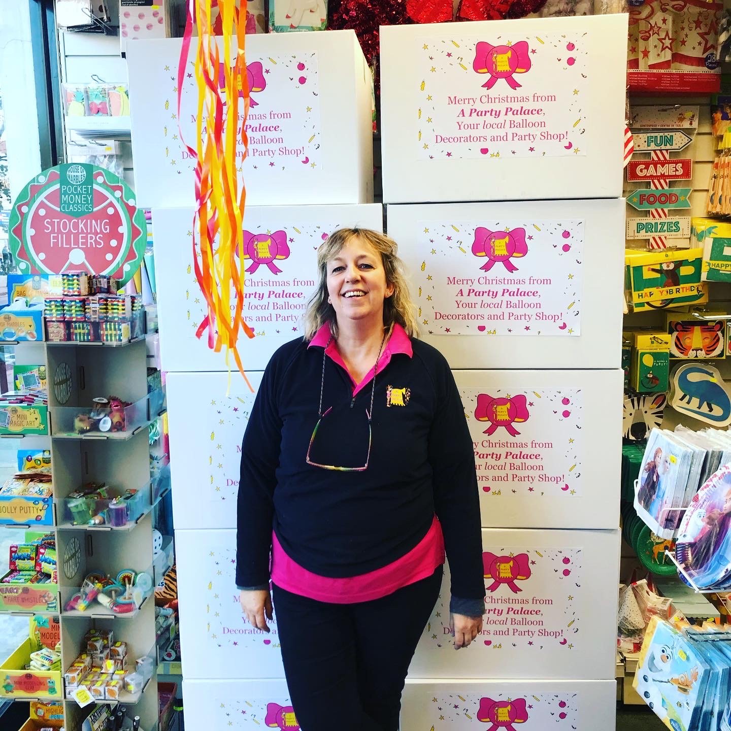 A woman smiling in a party supply store, standing in front of large boxes with Christmas decorations and balloons, with shelves of toys and party supplies on either side.