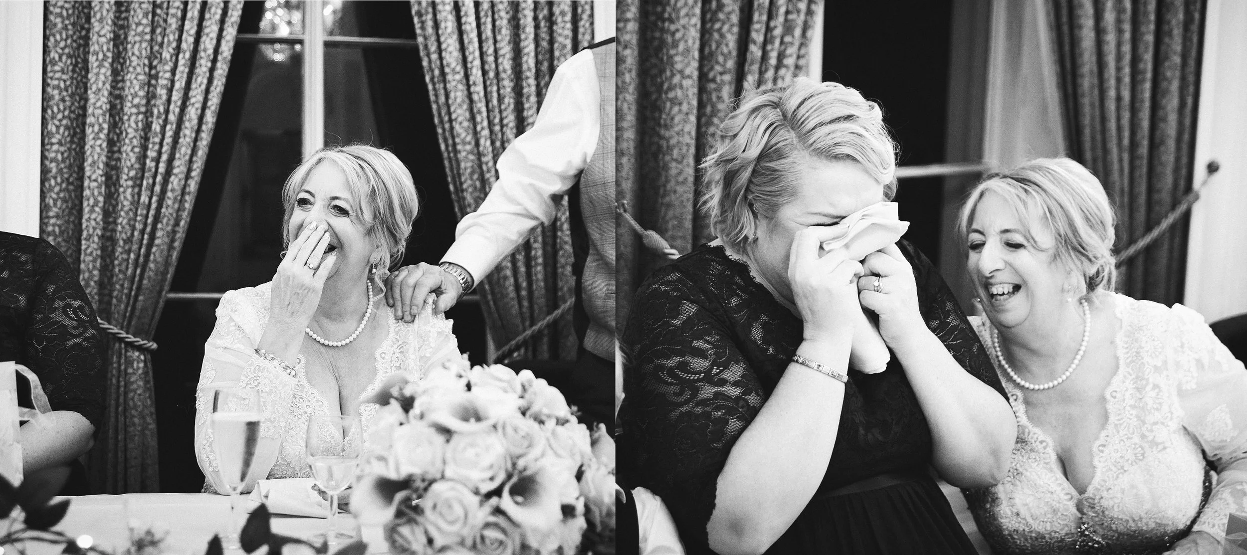 A woman in a wedding dress laughing and wiping away tears at a wedding reception, seated at a table with flowers and drinks, with a woman next to her also laughing.