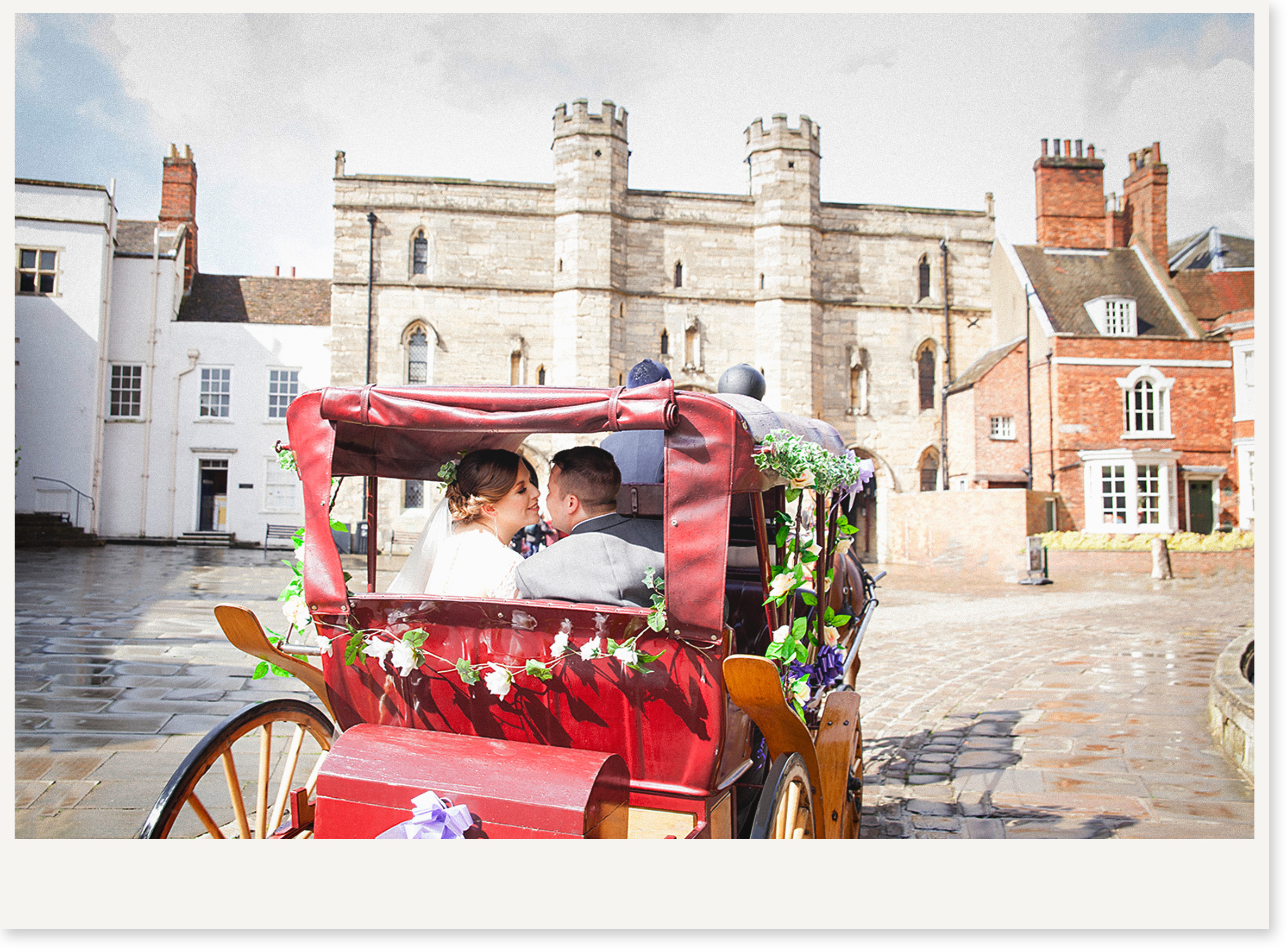 A couple riding in a red decorated horse-drawn carriage in a town square with historic buildings and a stone castle in the background.