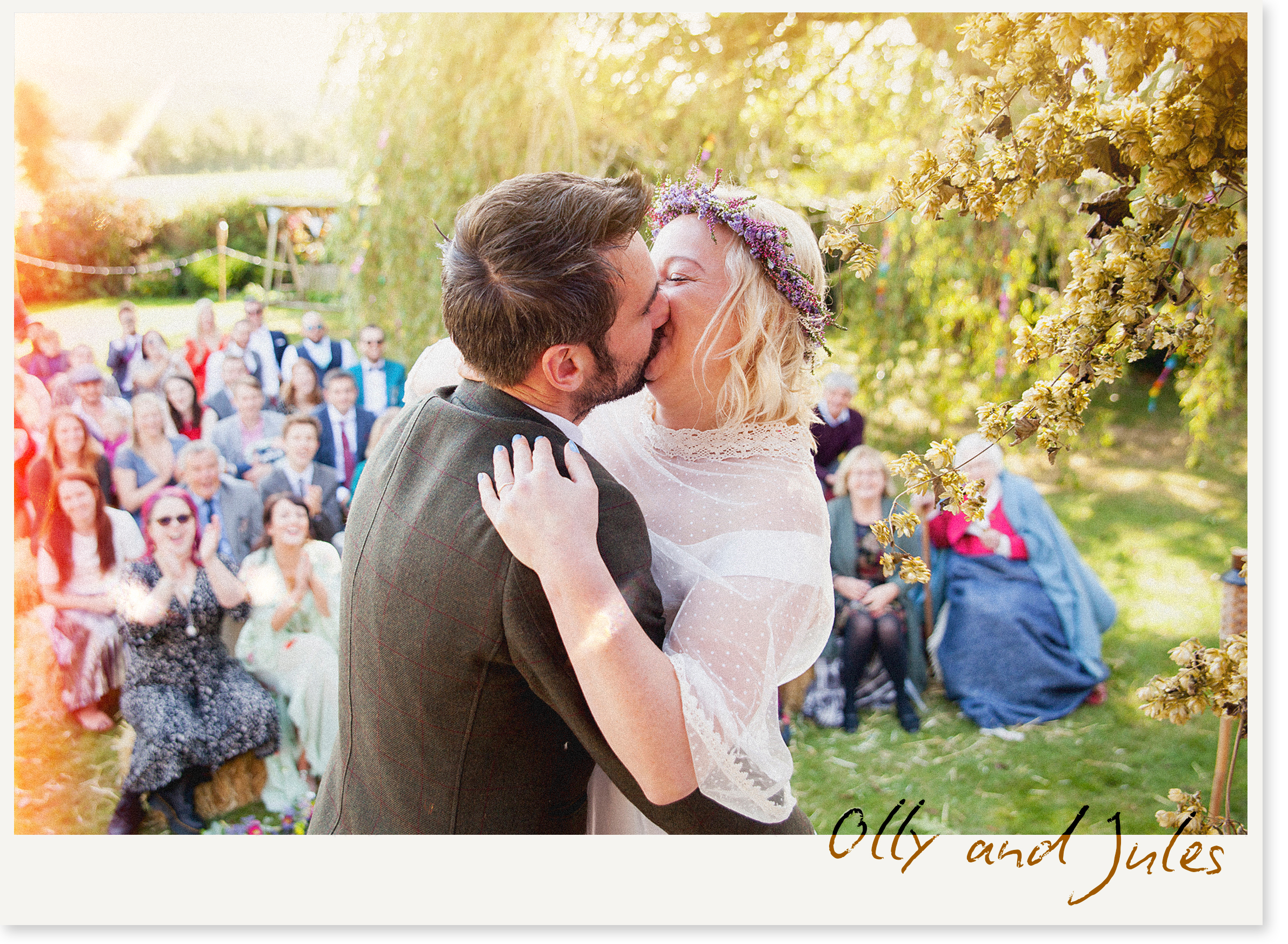 A bride and groom kiss during their outdoor wedding ceremony surrounded by friends and family, with yellow flowering trees and soft sunlight creating a romantic atmosphere.
