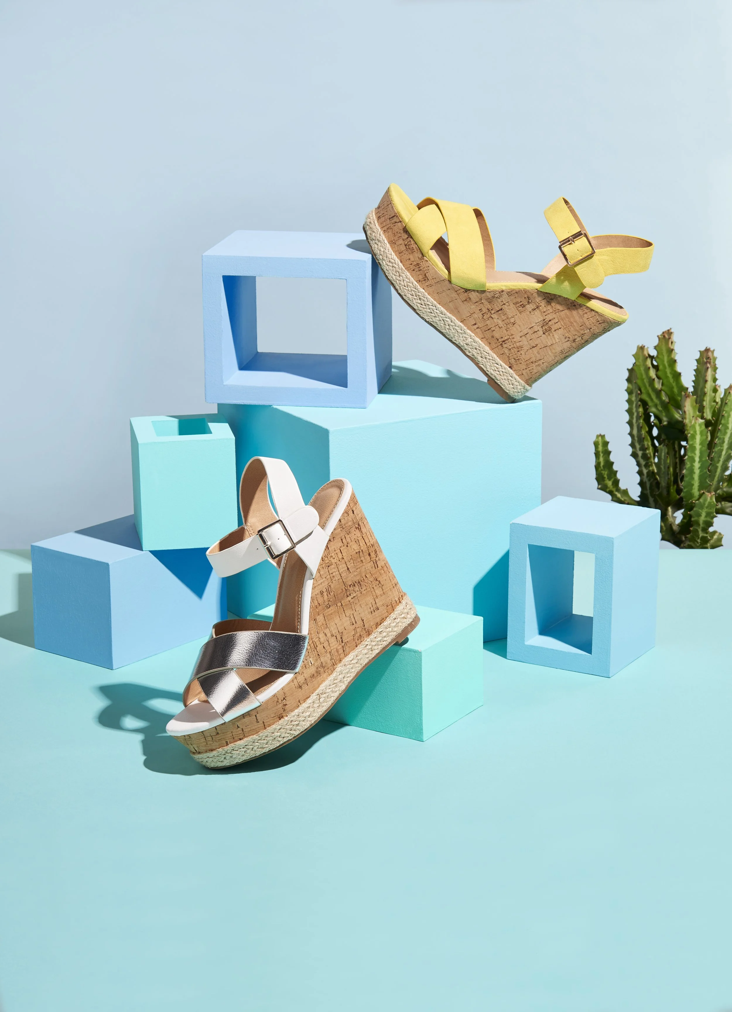 A pair of yellow platform wedge sandals and a pair of metallic silver and white sandals displayed on pastel blue platforms with geometric shapes and a green cactus plant in the background.