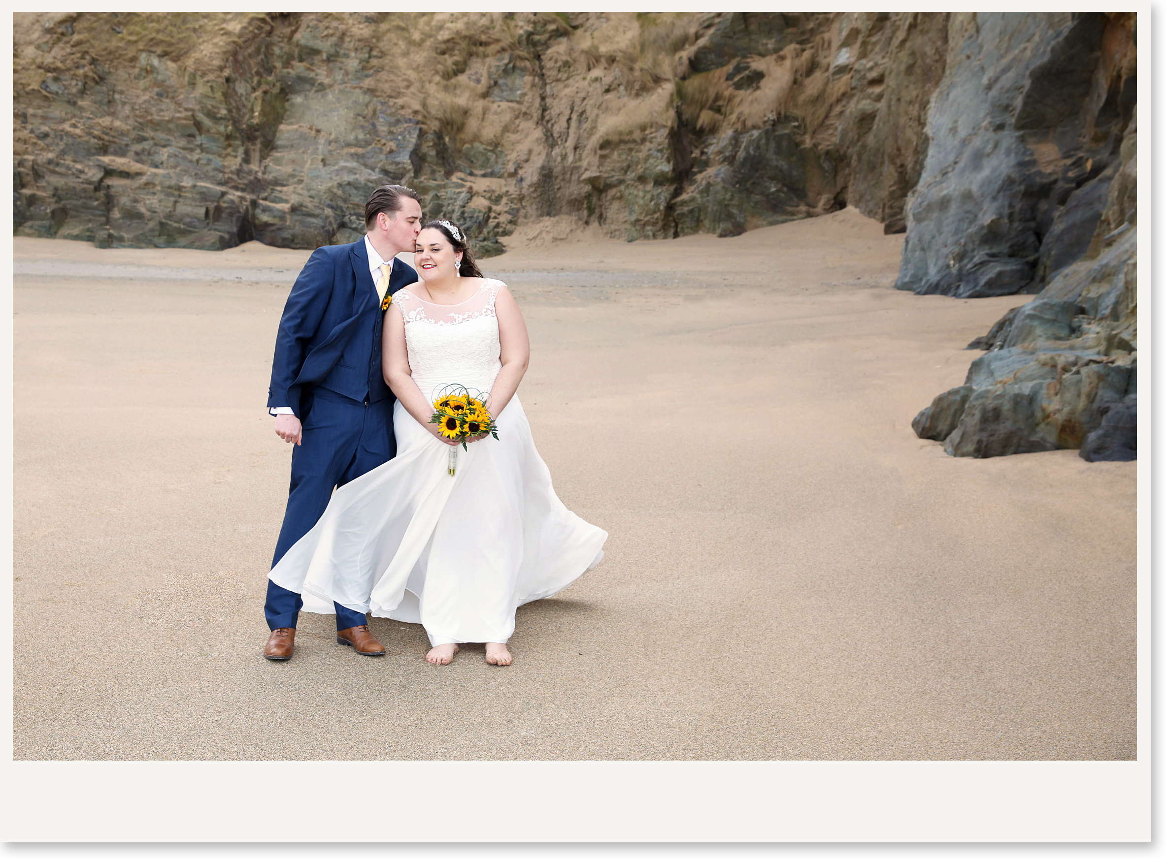 A newlywed couple on a sandy beach with rocky cliffs in the background. The groom is kissing the bride on the forehead, and the bride is smiling while holding a bouquet of sunflowers. The bride is barefoot, and the groom is wearing brown shoes.
