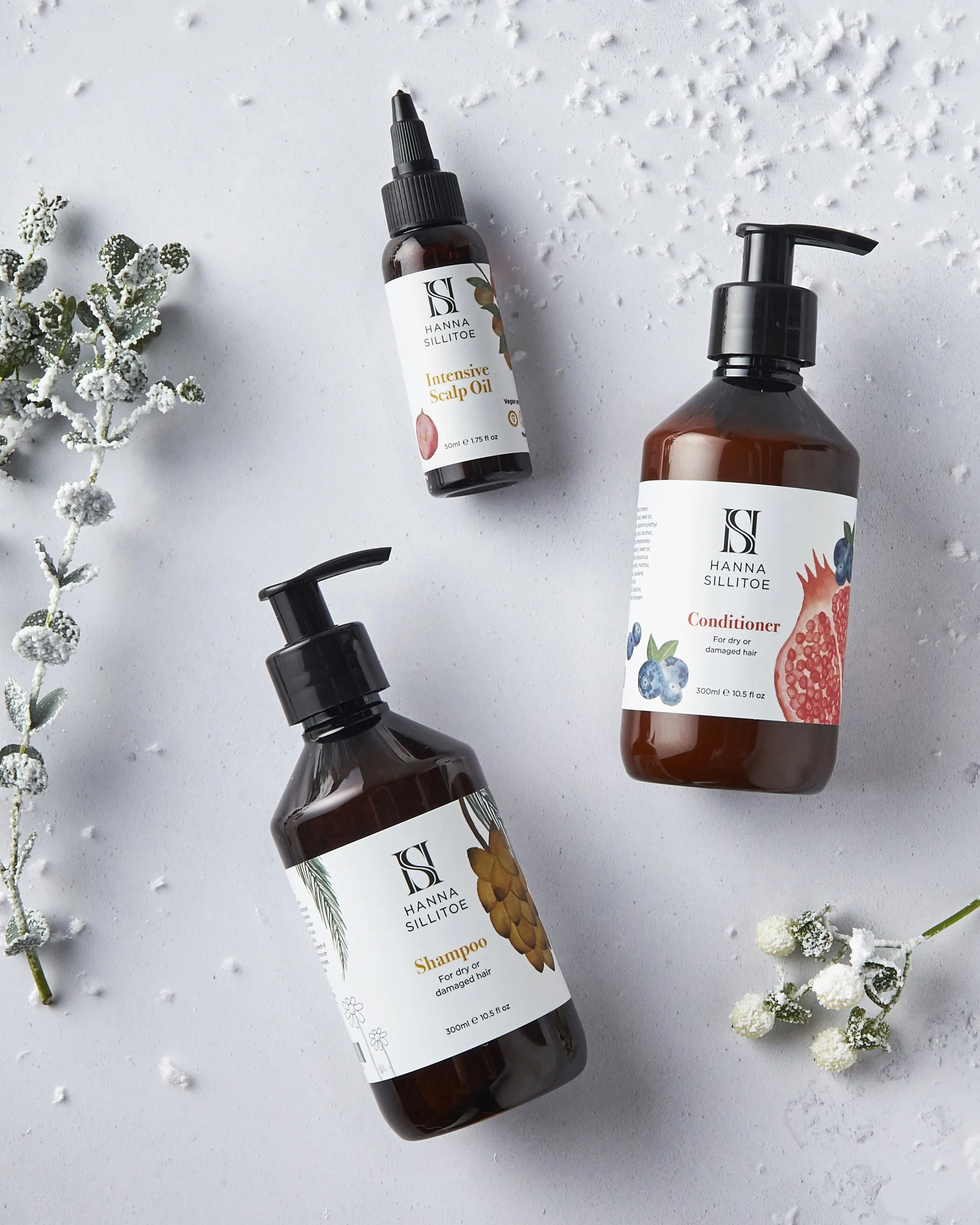Three bottles of Hanna Sillitoe hair care products—shampoo, conditioner, and intensive scalp oil—are arranged on a white textured surface with small white flowers and a sprig of greenery around them.