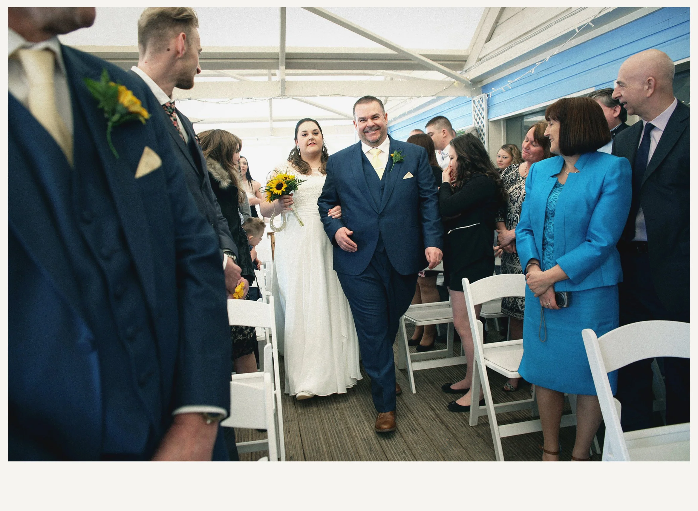 A wedding reception with guests standing and smiling. The bride, holding a sunflower bouquet, is walking down an aisle. The groom and guests are dressed in formal suits and dresses, engaging in conversations and looking happy. The setting is an indoo