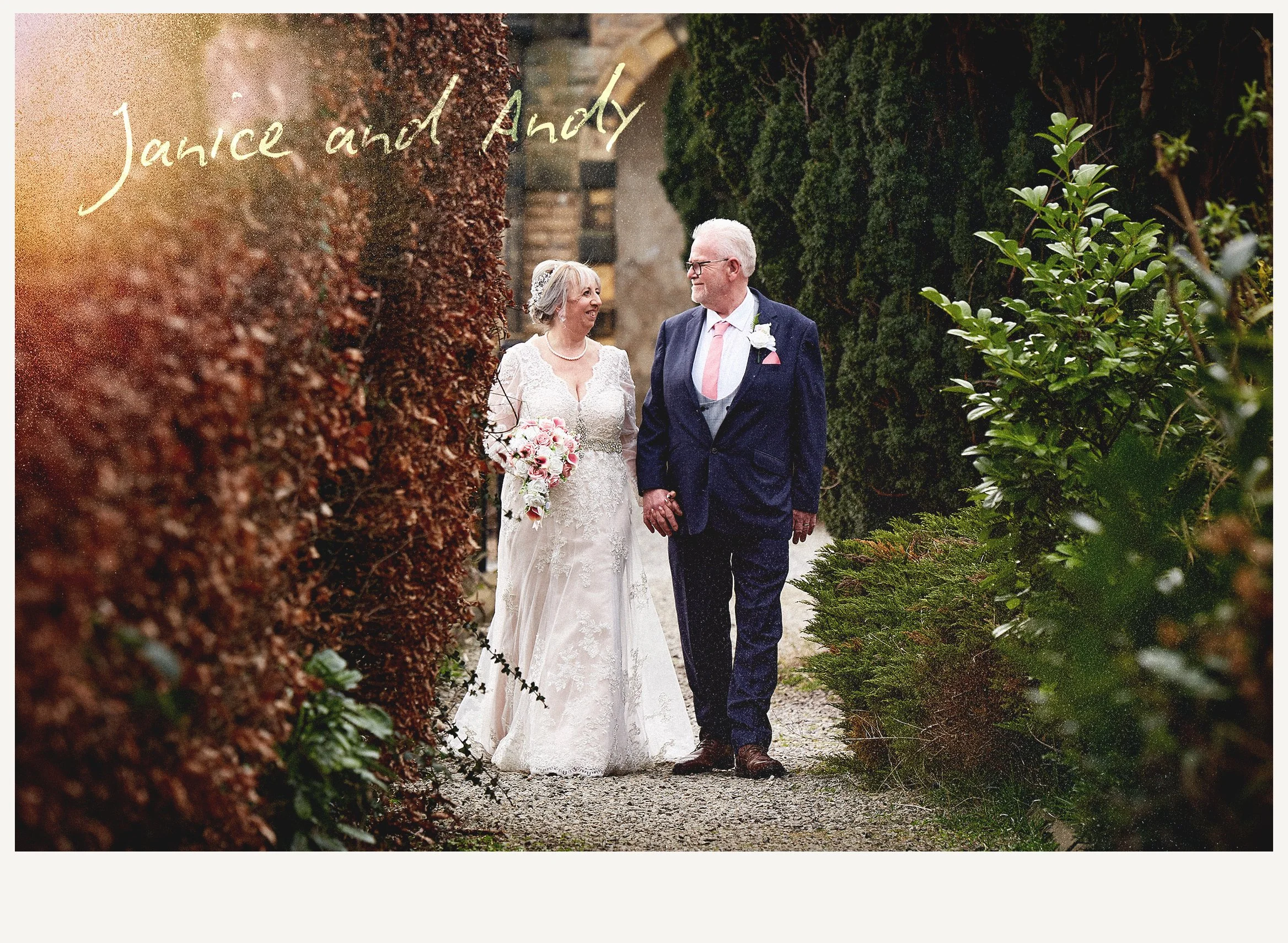 A bride and groom walking outdoors on a gravel path, holding hands, surrounded by green bushes and trees. The bride wears a white wedding dress and holds a bouquet of pink and white flowers, while the groom wears a dark suit with a pink tie.