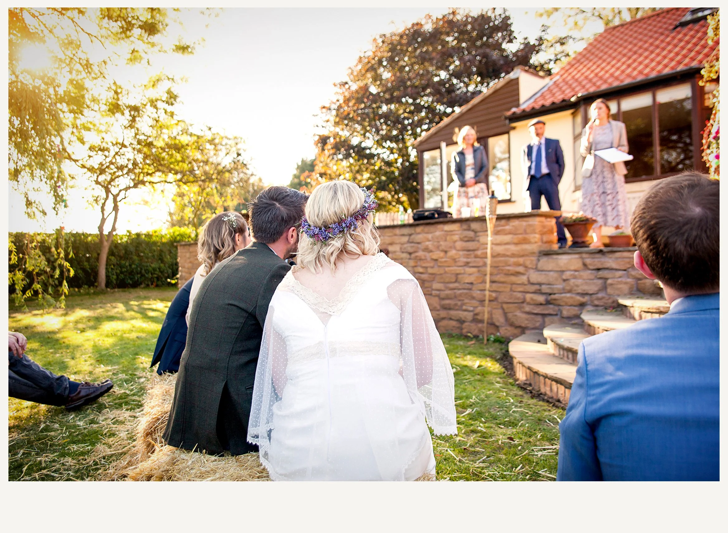 A couple dressed in wedding attire kneeling on a hay bale at an outdoor wedding ceremony, facing a small group of guests with a person speaking at a makeshift podium in front of a house with large windows and a red tiled roof, during sunset on a sunn