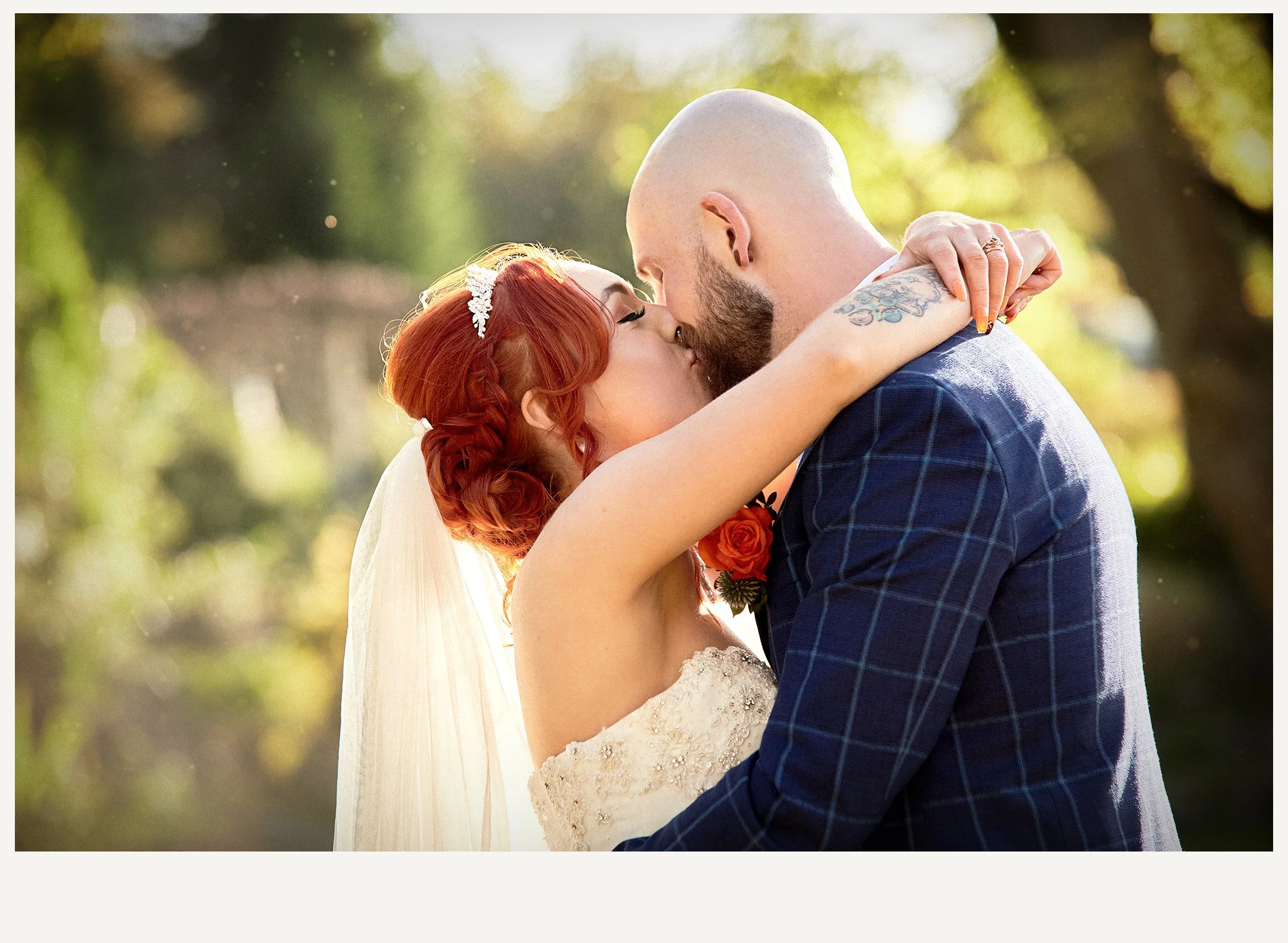 A bride and groom are embracing and kissing outdoors in a natural setting, with blurred green foliage and sunlight in the background.