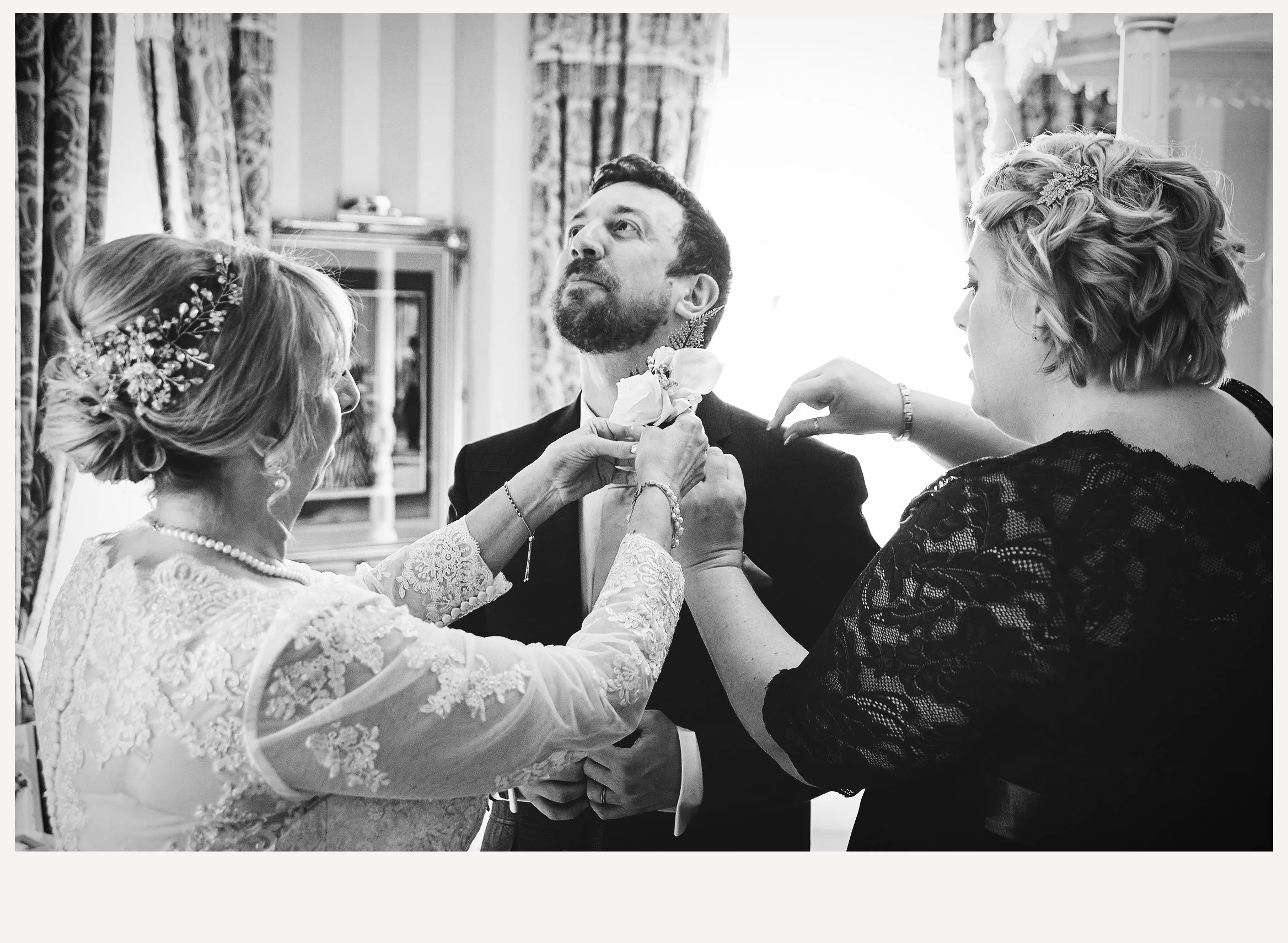 A black and white photo of a wedding scene where an older woman is adjusting the boutonniere of a groom, with two women on either side, one with curly hair and a lace dress, and the other with braided hair and a floral hairpiece.
