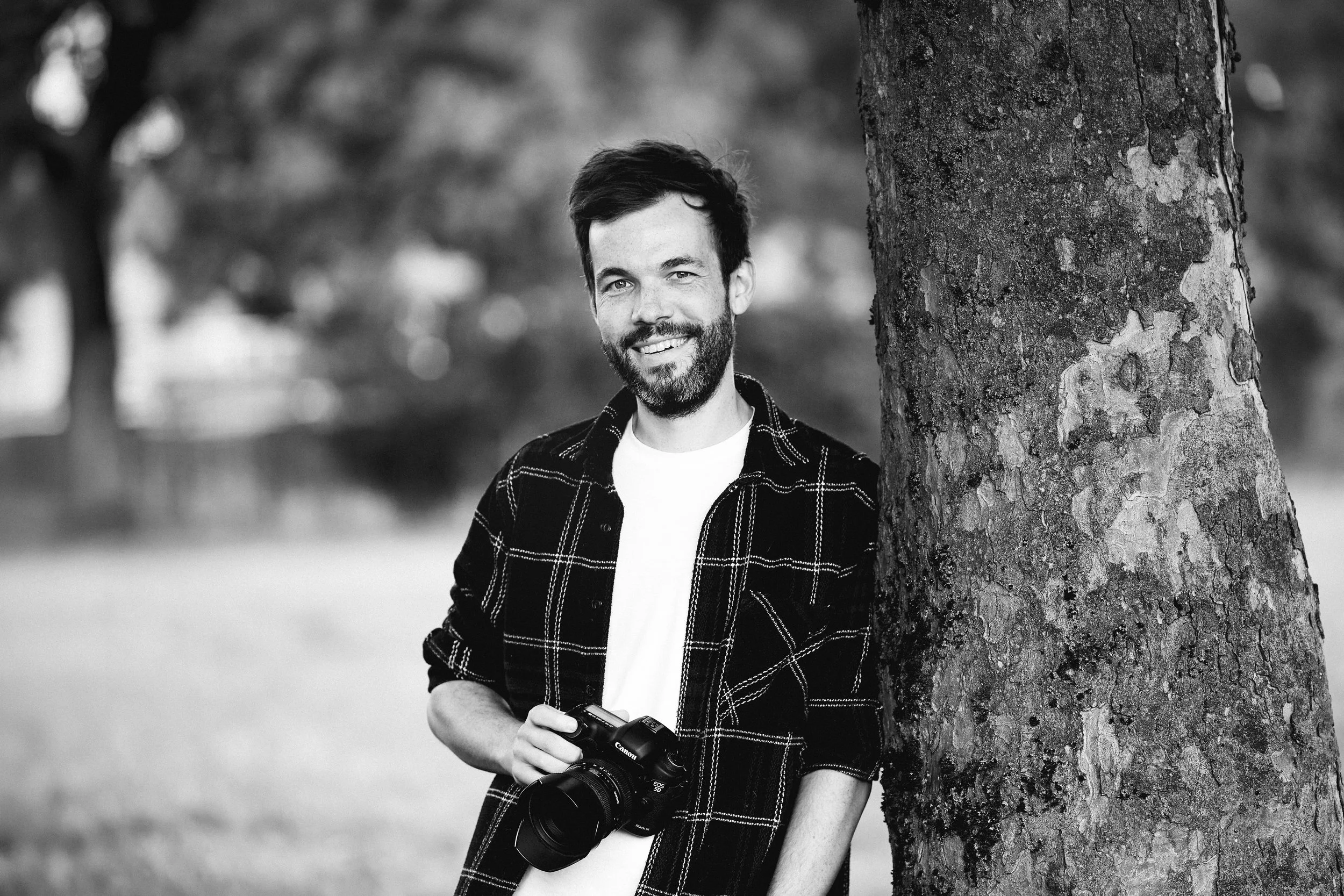 A smiling man with dark hair and a beard stands outdoors next to a tree, holding a DSLR camera. He is wearing a white shirt and a dark checked shirt. The background is blurred, showing more trees and open space in black and white.