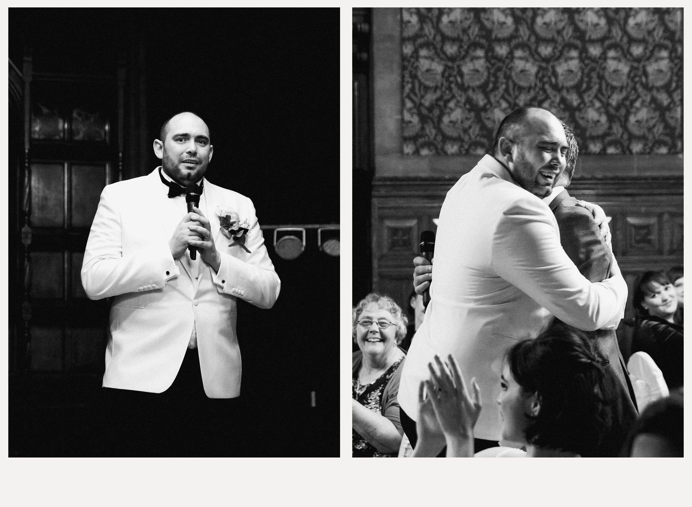 Black and white photo of a man in a tuxedo holding a microphone, speaking at an event. In the background, there are people seated, some smiling, with one woman clapping and a man hugging the speaker.
