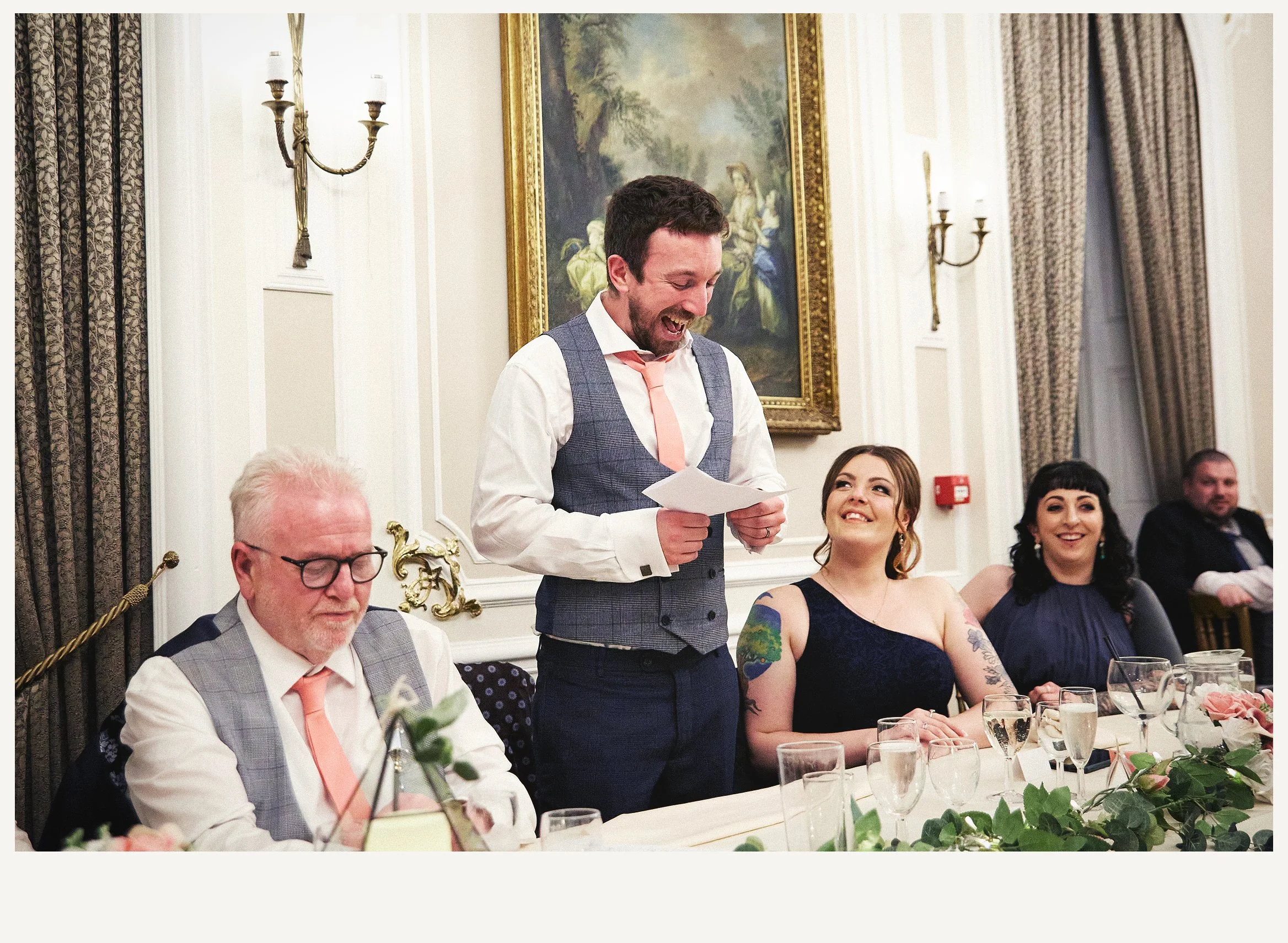 A man in a vest and tie is giving a speech at a formal dinner event, with several other seated guests in an elegant room with framed painting and ornate decor.
