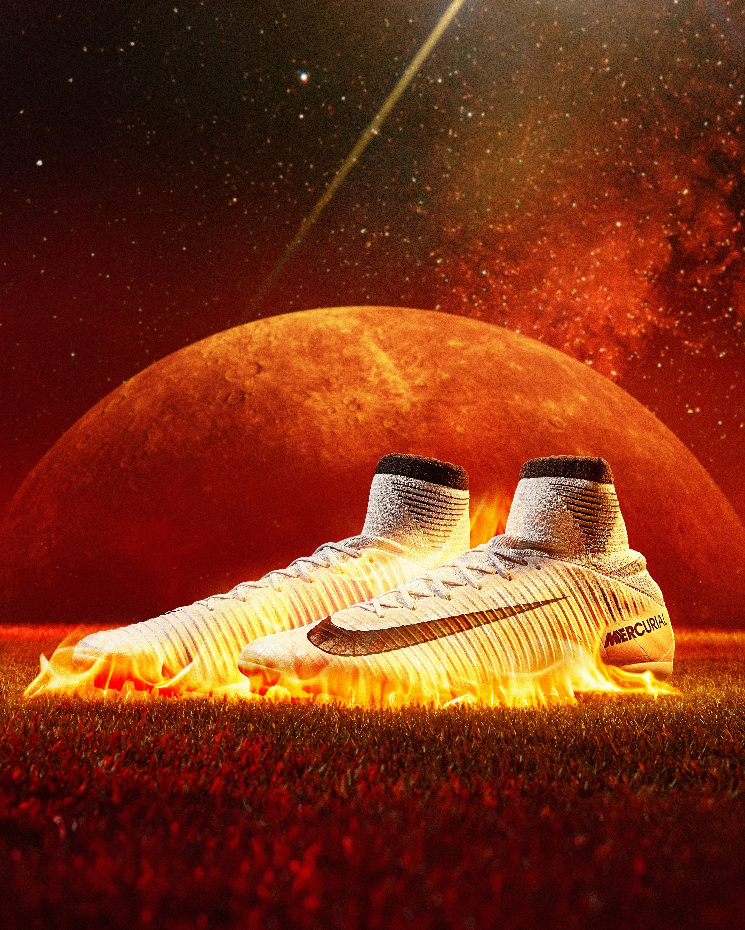 White Nike Mercurial soccer cleats with black and gold accents and black Nike logo, lying on the ground with flames beneath, with a red planet and starry space in the background.