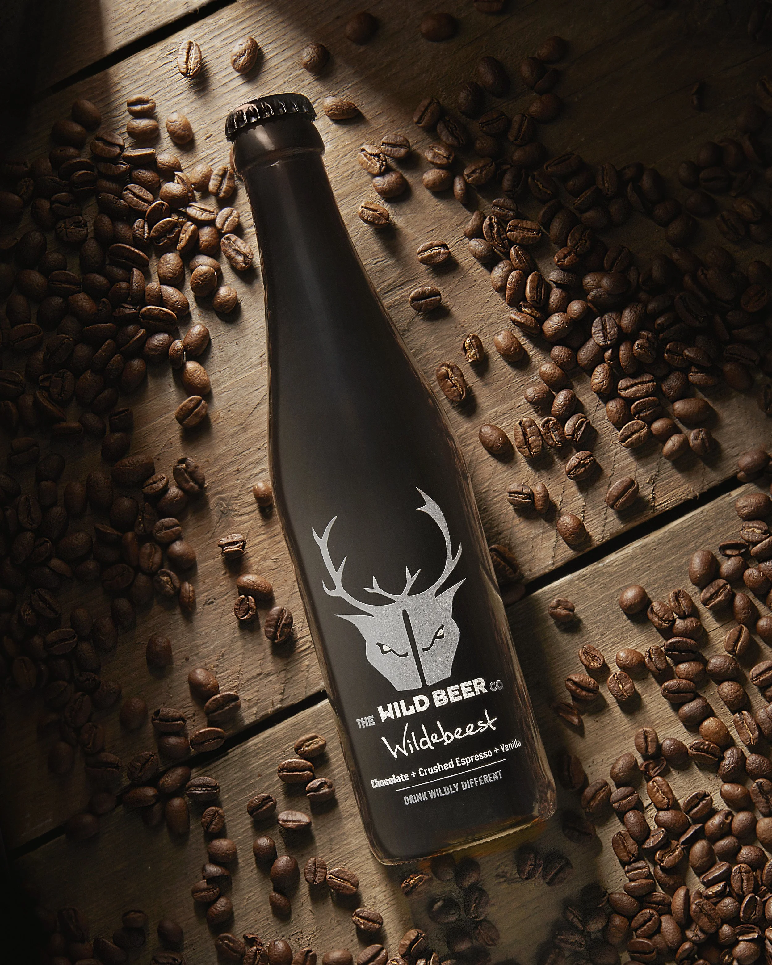 A black bottle labeled 'The Wild Beer Co.' with a white deer skull logo, lying on a wooden surface surrounded by scattered coffee beans.