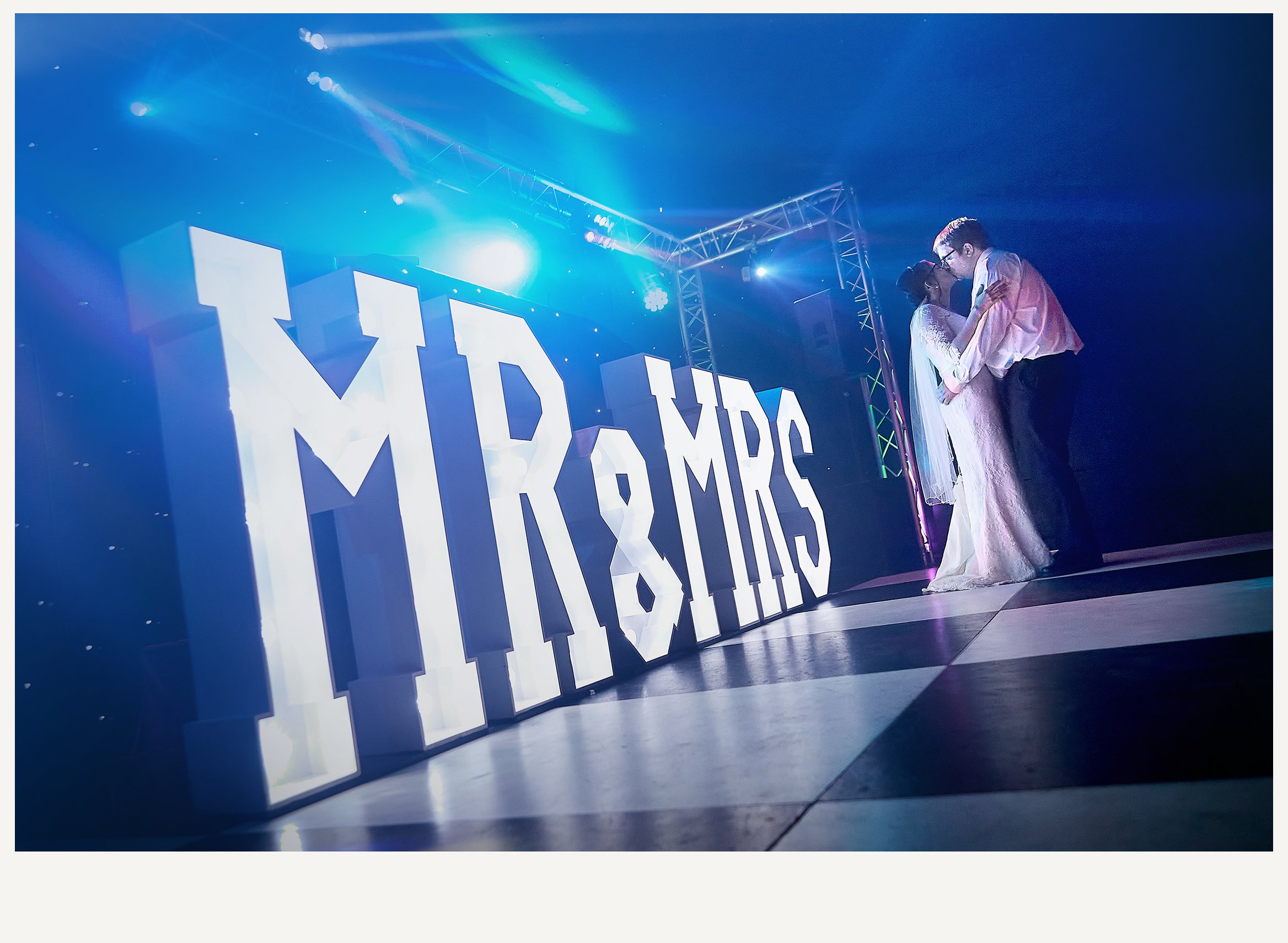 A large illuminated sign with the word 'MAYBACH' in bold white letters against a dark background, with two people, a man and a woman, dressed in formal attire, sharing an intimate moment at an event. The scene is lit with blue and purple lighting.