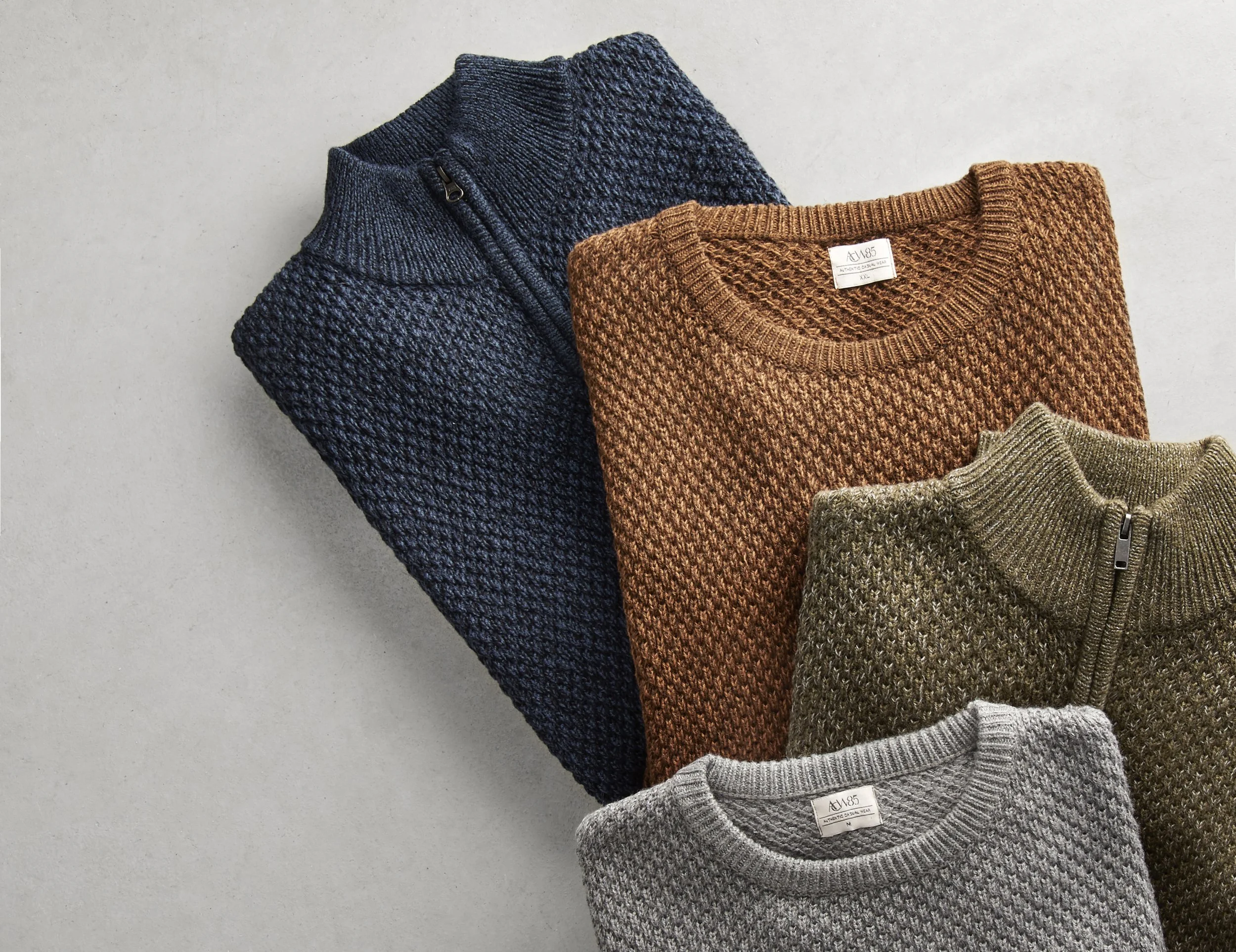 Four folded sweaters in dark blue, brown, gray, and beige colors laid on a light surface.