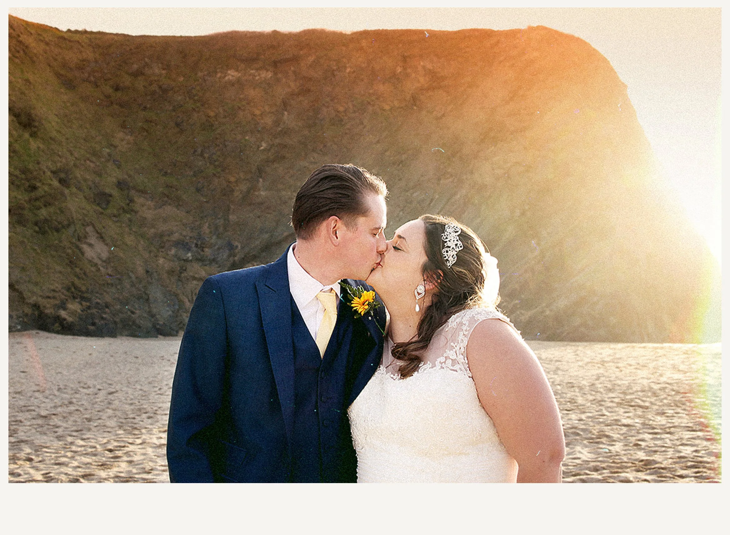 A bride and groom kissing on the beach during sunset. The groom is wearing a blue suit and the bride is wearing a white lace wedding dress with a floral headpiece.