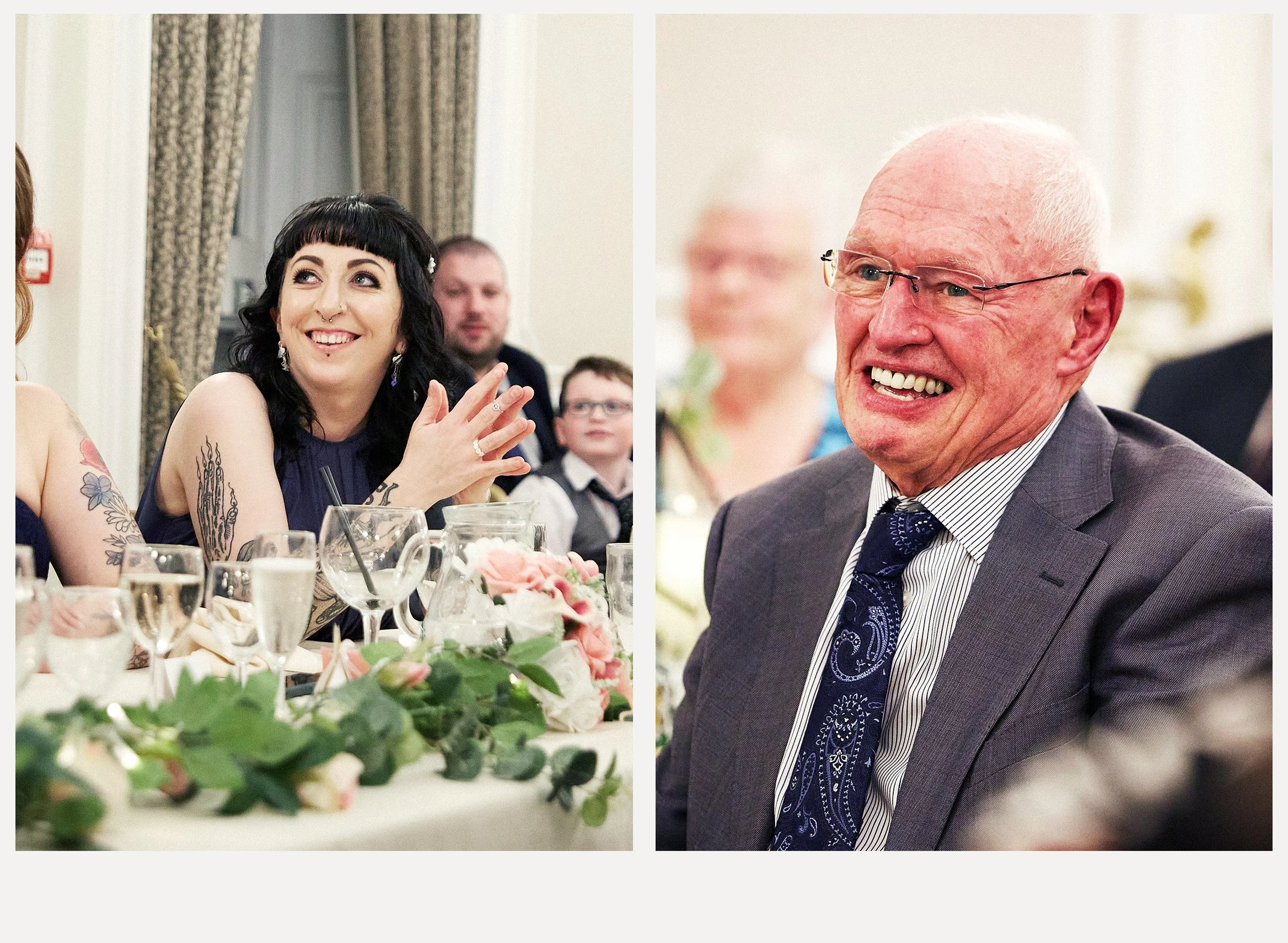A woman with dark hair, tattoos, and earrings smiling and clapping at a formal event, seated at a table decorated with flowers and glassware. To her right, a bald man with glasses wearing a gray suit and patterned tie also smiling. In the background,