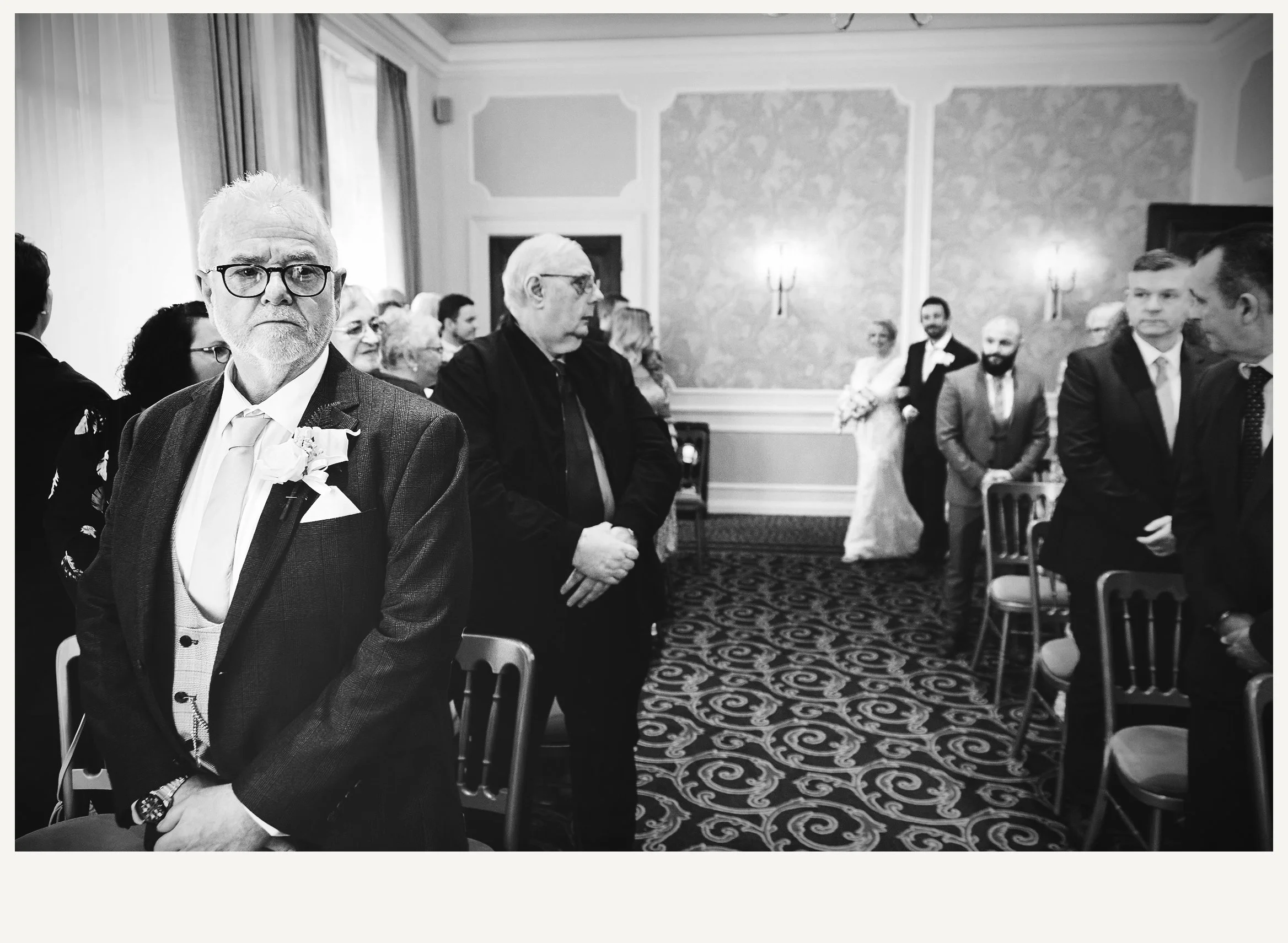 Black and white photo of people attending a formal event in a decorated room, with chairs arranged in rows and a patterned carpet.