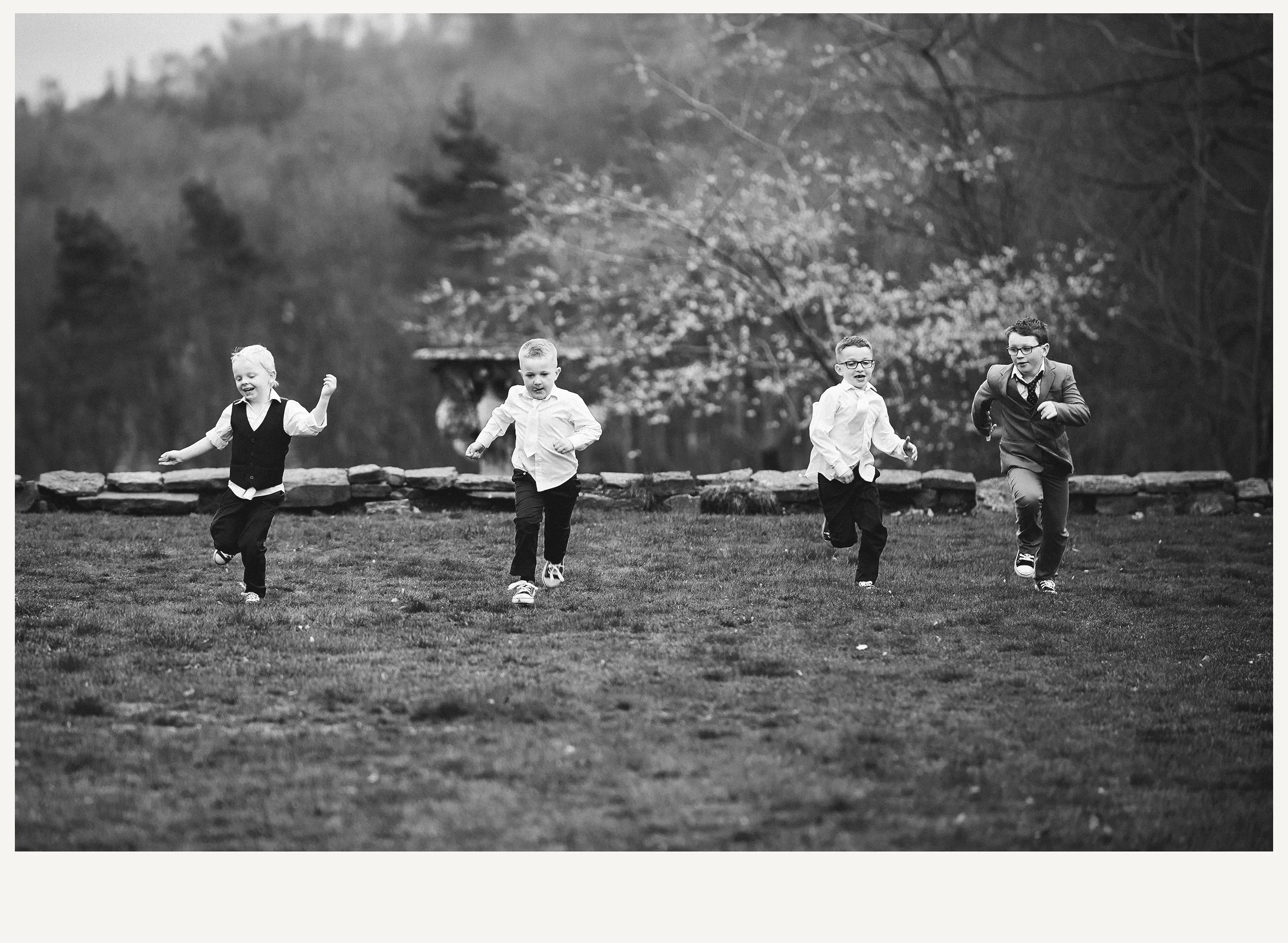 Four young boys running and playing outdoors on a grassy area with trees and a stone fence in the background.