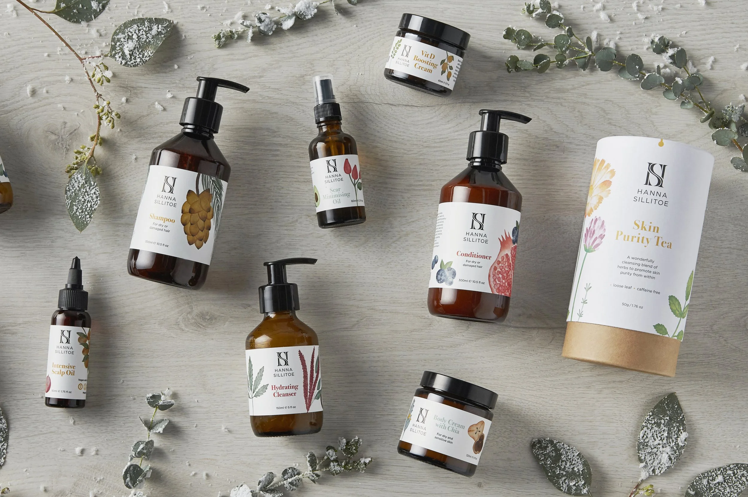 Flat lay of skincare and haircare products on a light wooden surface, including bottles, jars, and a tube, accompanied by sprigs of eucalyptus and decorative green leaves.