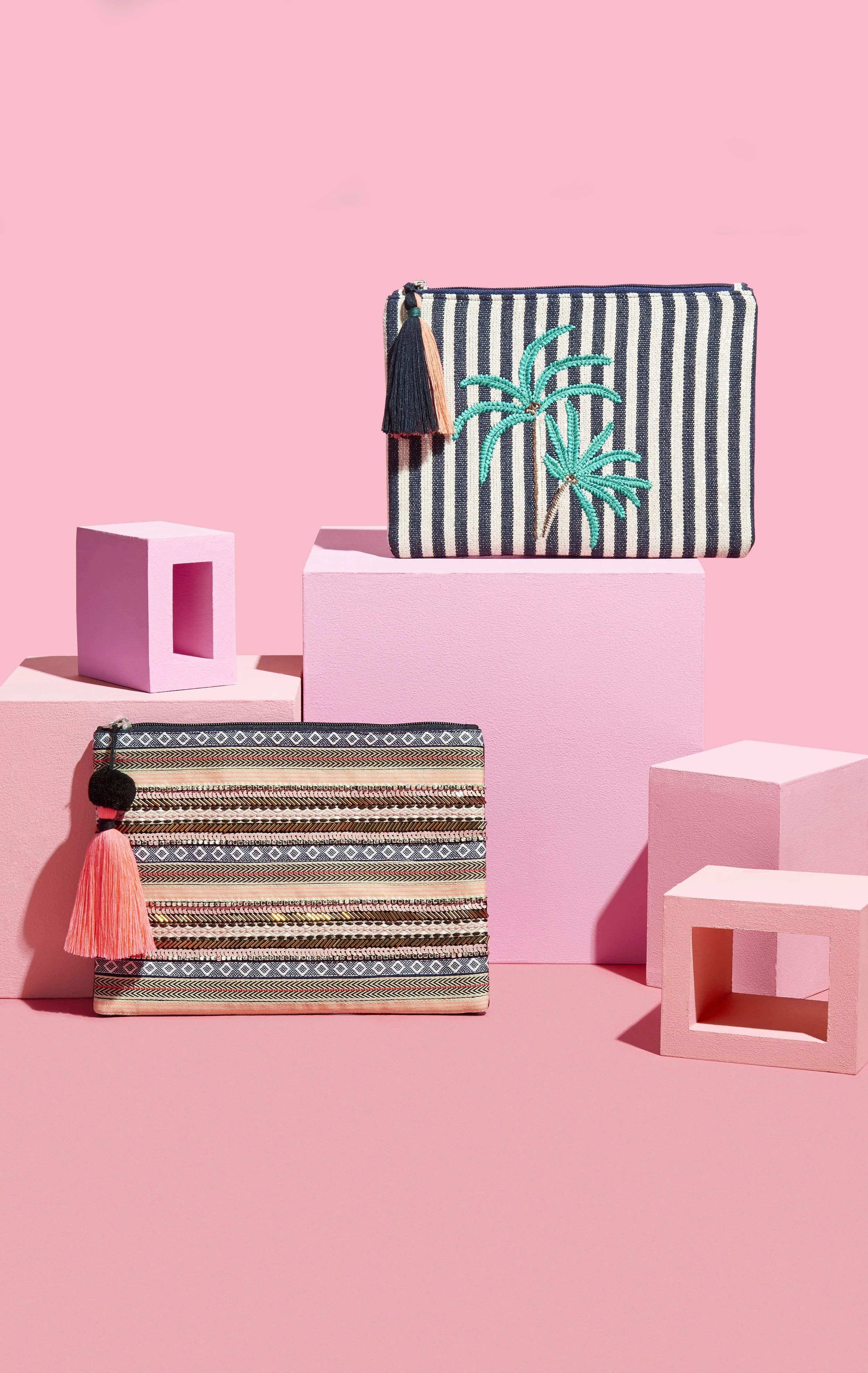 Two woven clutch purses with tassels and embroidered palm trees, displayed on pink geometric pedestals against a pink background.