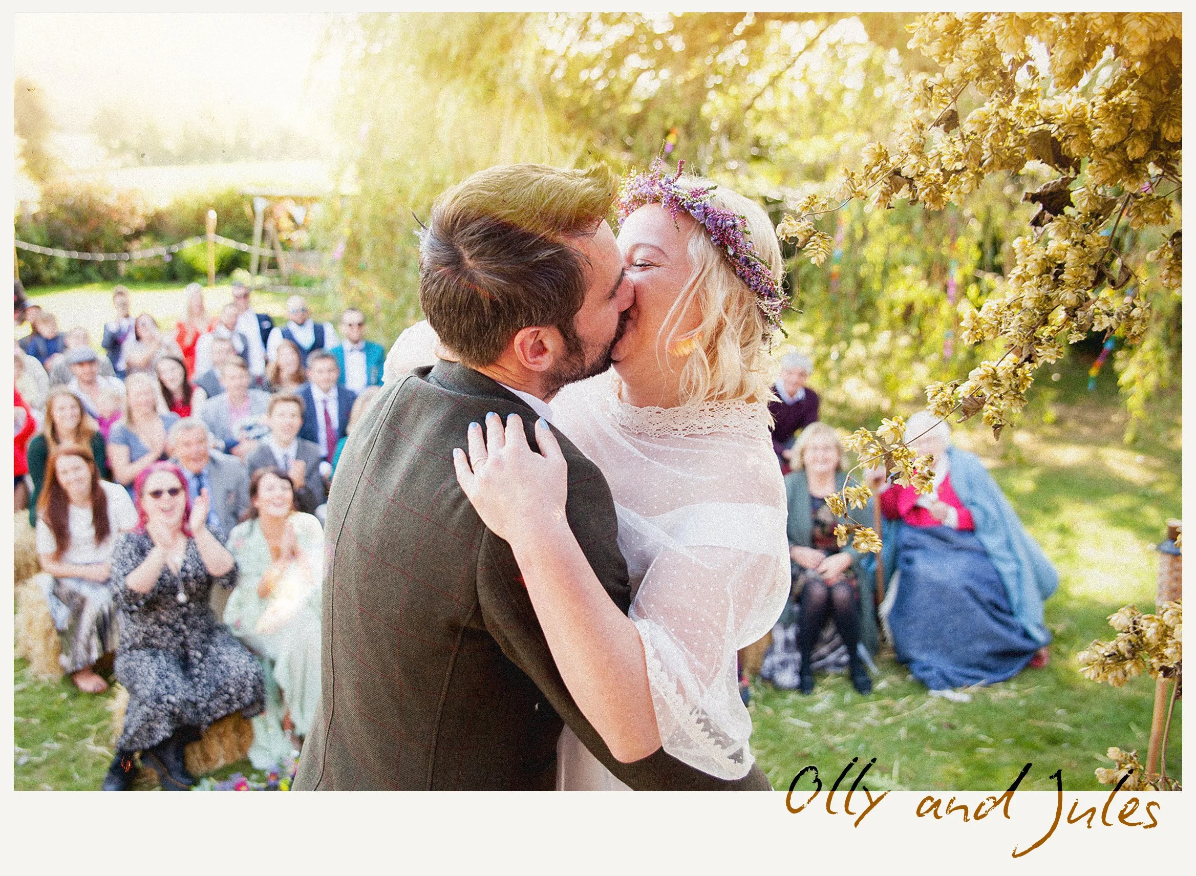 A couple, dressed in wedding attire, sharing a kiss outdoors during their wedding ceremony, with a group of seated guests in the background and trees with yellow leaves framing the scene.