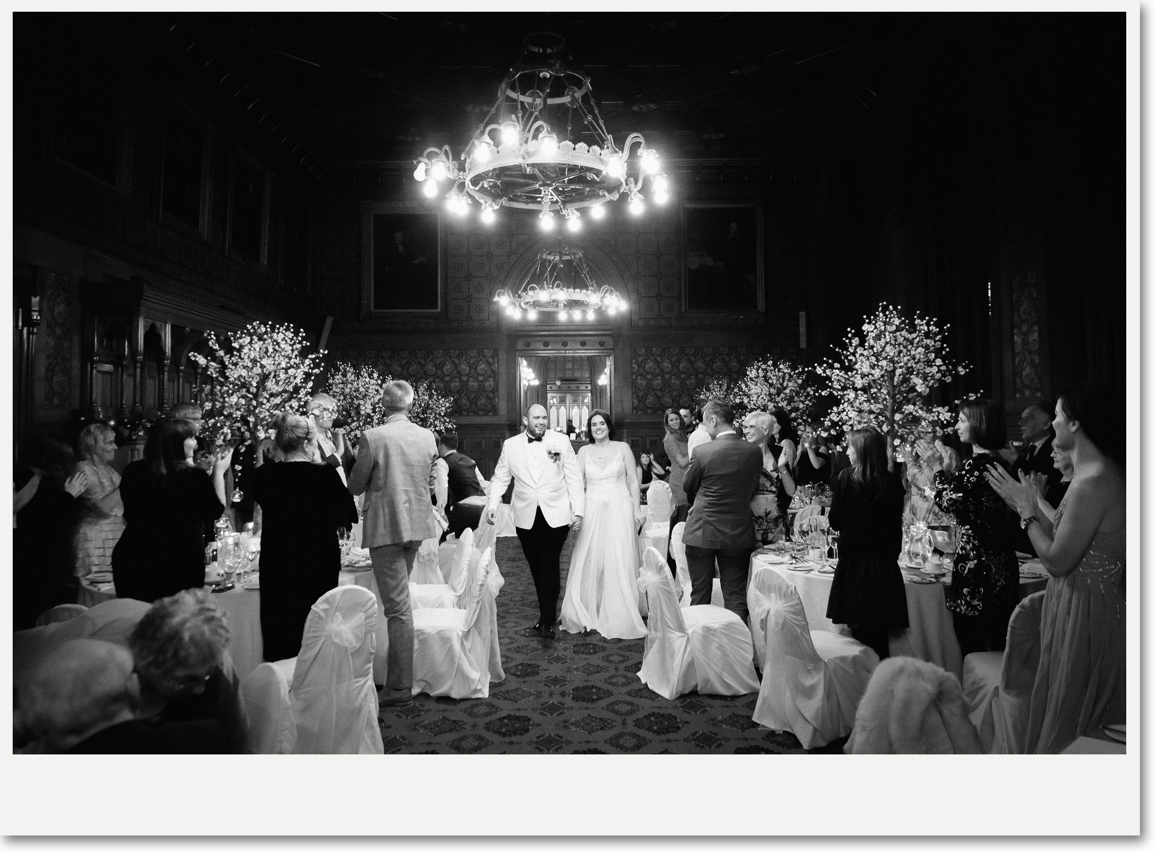 A wedding reception with a bride and groom walking through a room filled with guests. The room is decorated with chandeliers and floral arrangements, with guests clapping and smiling.