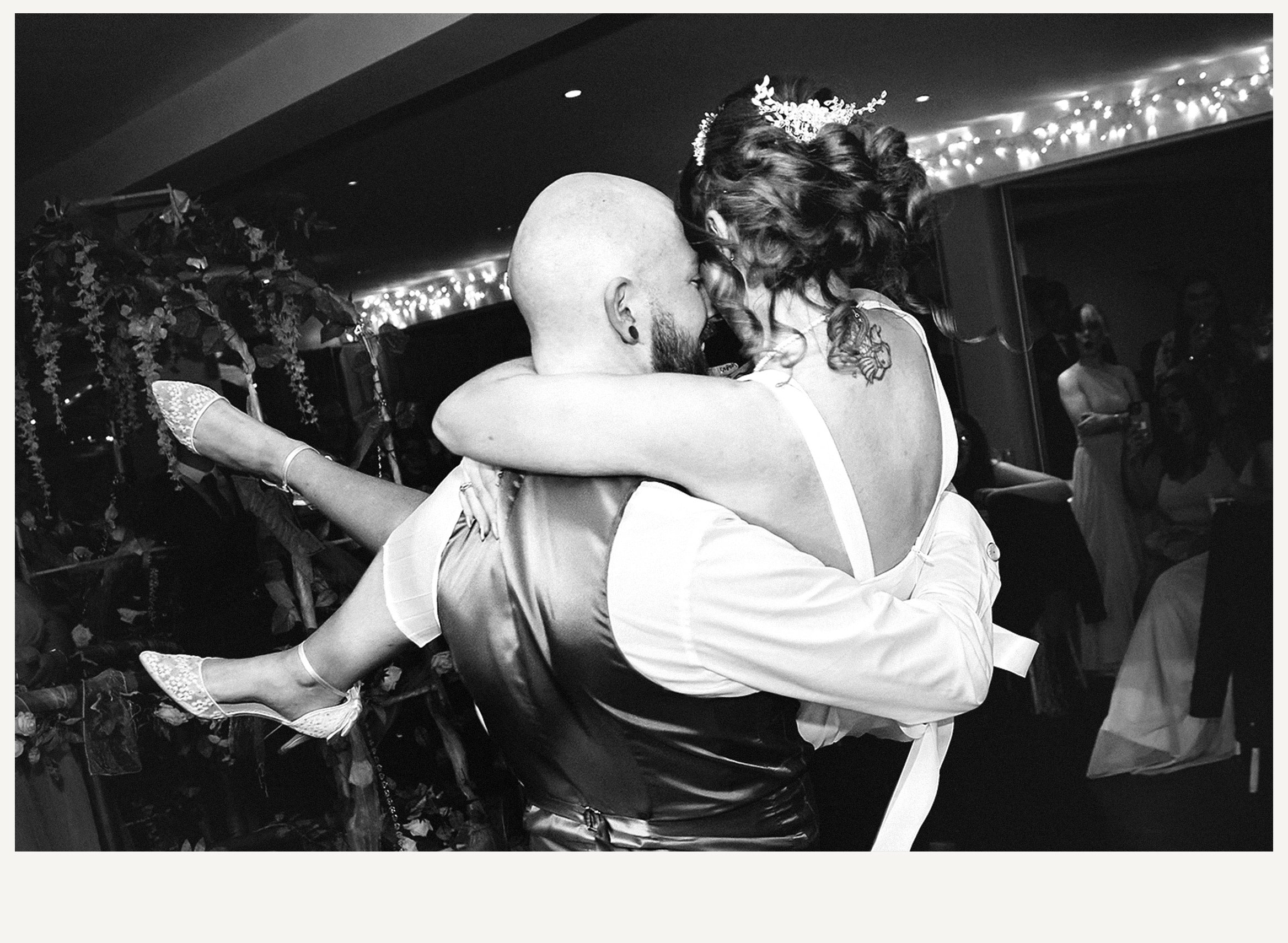A couple is dancing closely at a celebration, with the man bald and the woman with curly hair. They are embracing, and the woman is wearing lace shoes. The background is decorated with string lights and floral arrangements.