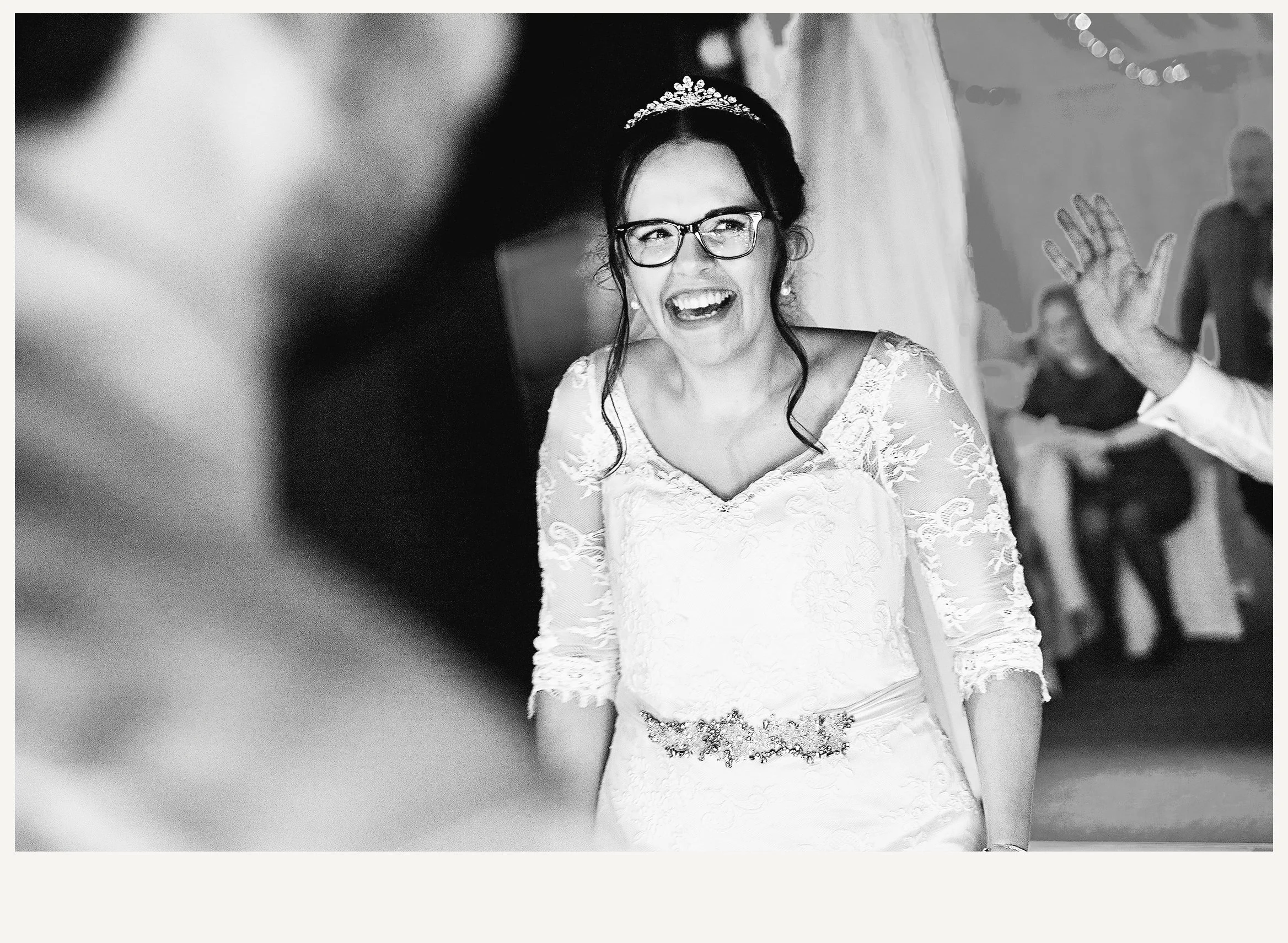 A woman in a wedding dress wearing glasses and a tiara, smiling with her mouth open, waving her left hand, in a black and white photo.