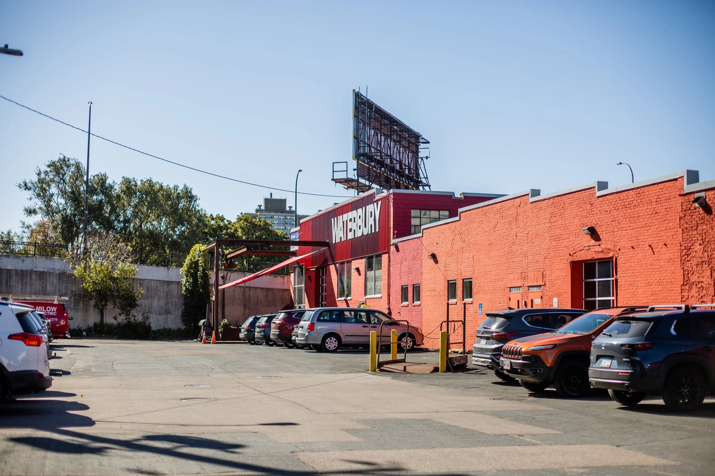 Parking lot with several cars parked in front of a red brick building. The building has a large sign that reads 'Waterbury'. There are trees with autumn foliage and a billboard structure above the building.