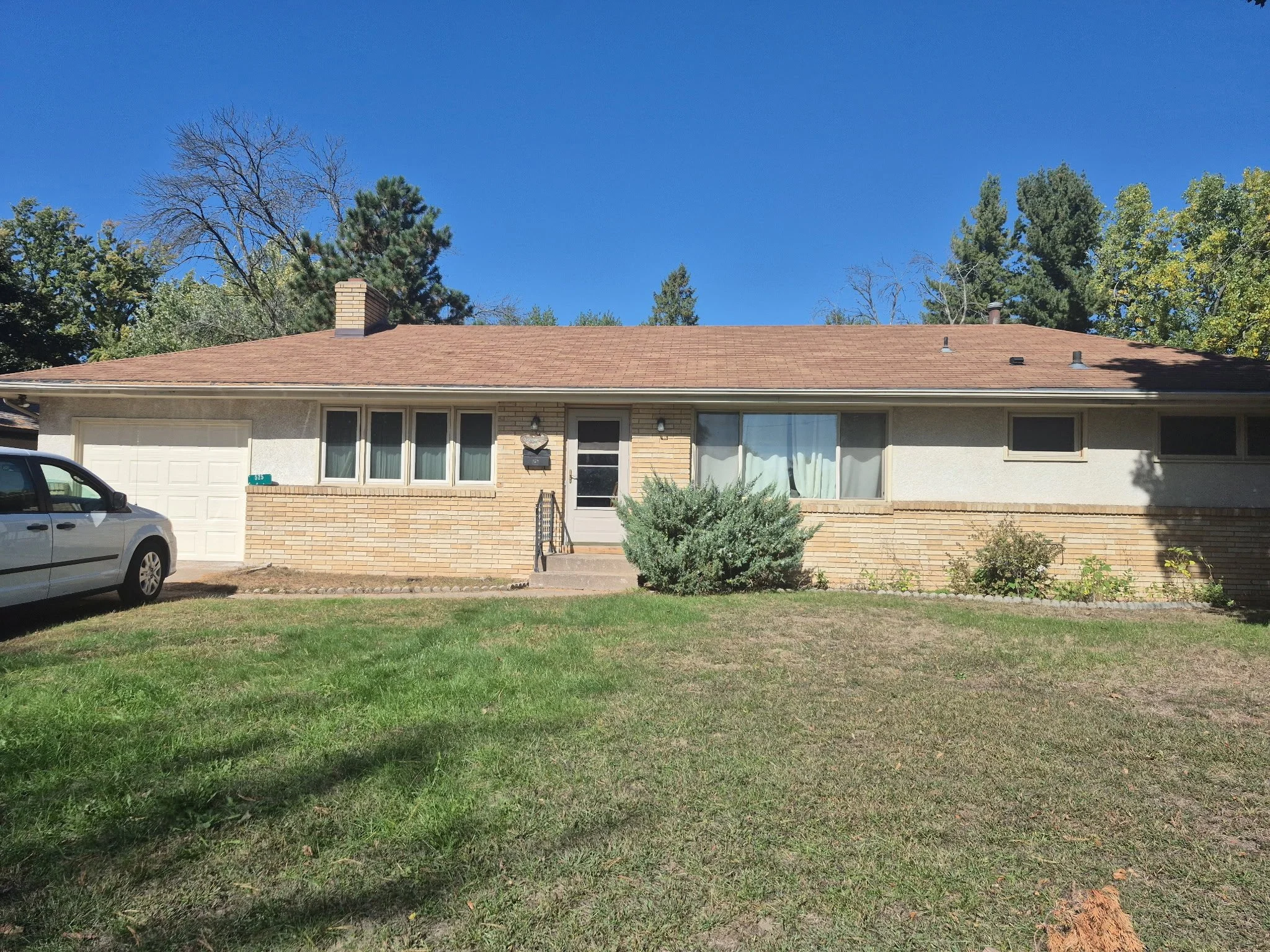 A house with a reddish-brown roof, brick and stucco exterior, front windows, and a small front yard with green grass and bushes. A driveway with a white and black car parked on it is visible on the left.