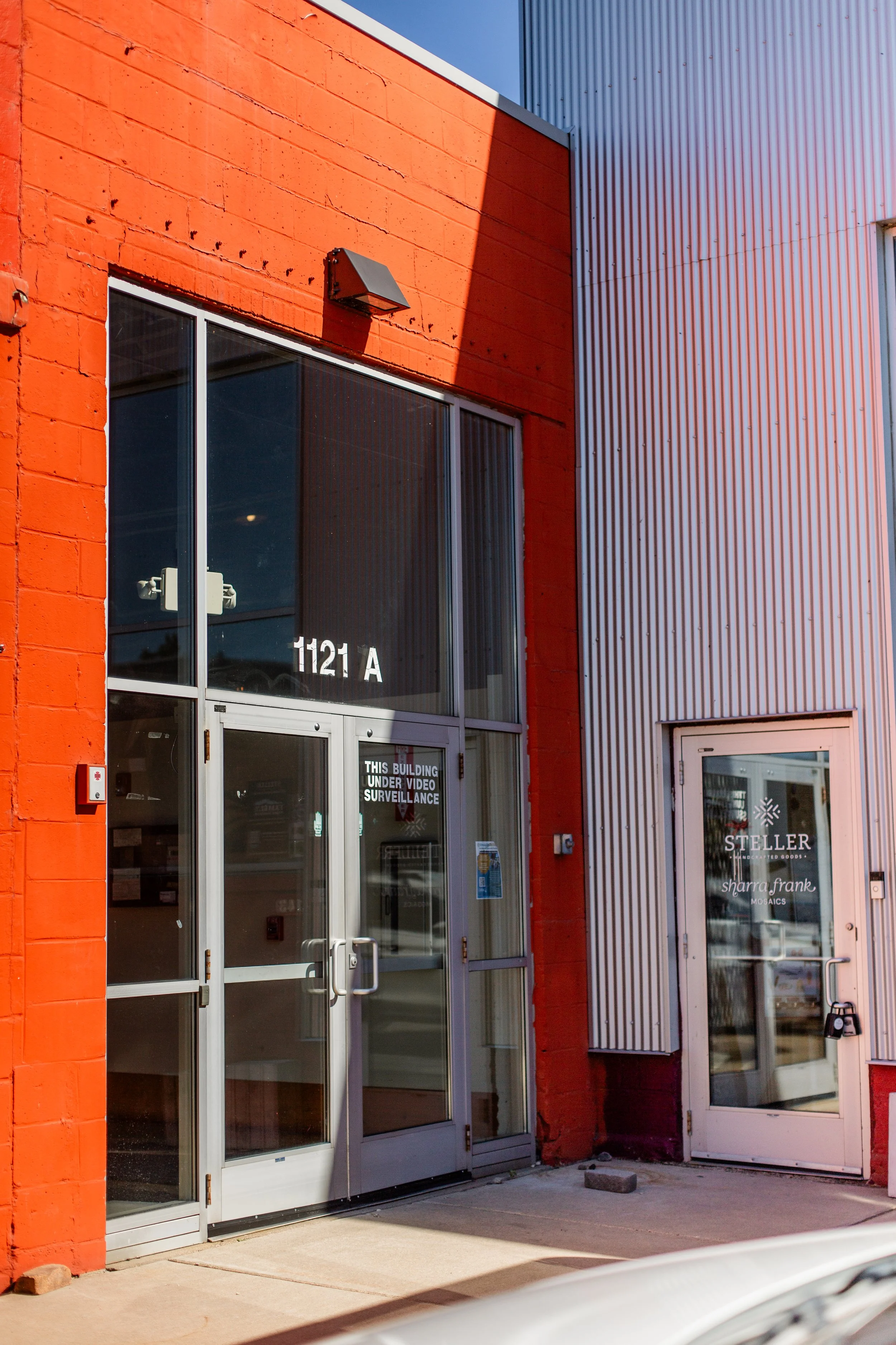 The entrance to a commercial building with glass doors, displaying the street number 1121A, and a sign that says 'This building under video surveillance'. The building has a red-painted brick wall and a adjacent metal panel wall, with a small securit