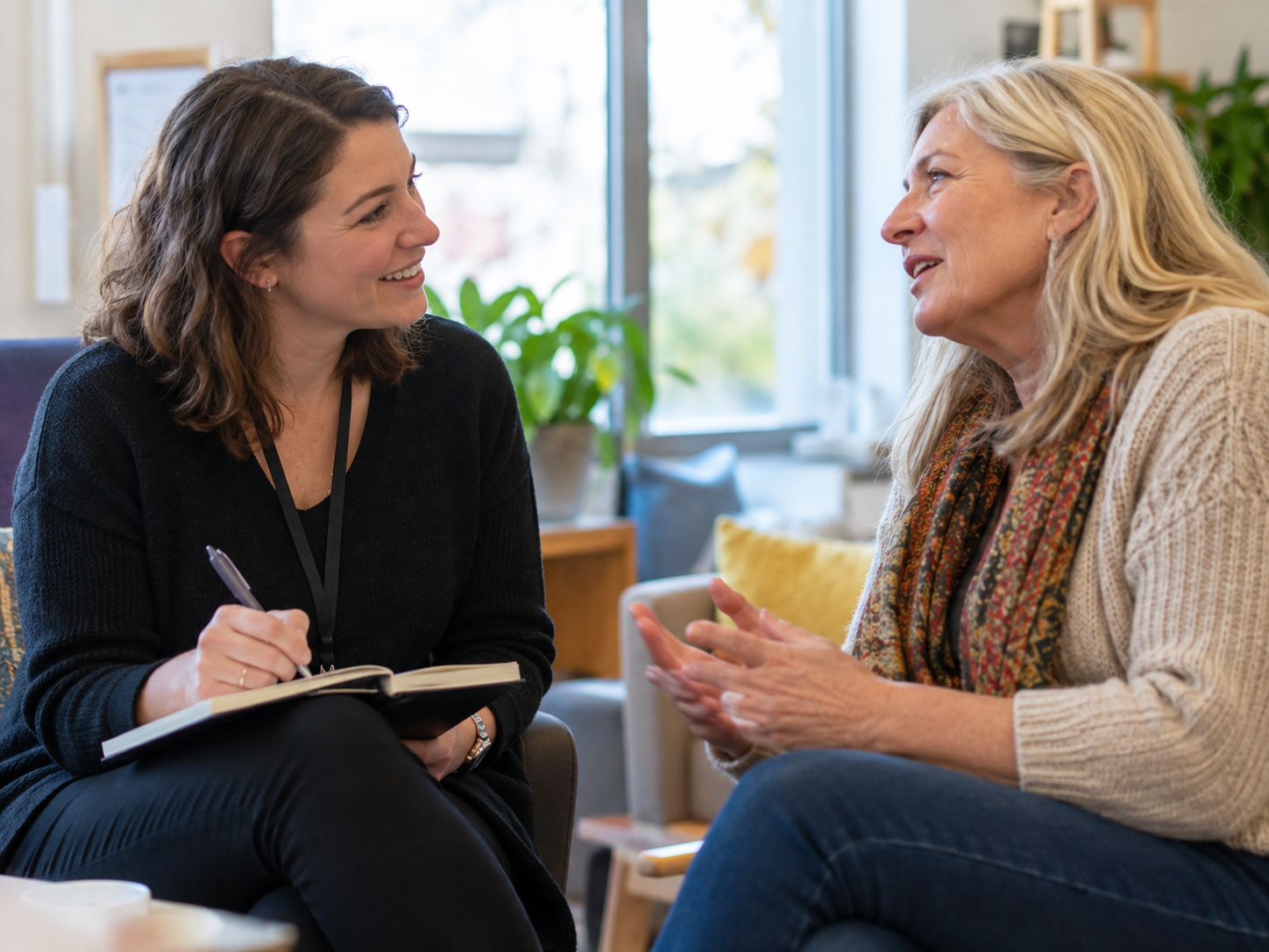 Two women talking in a living room, one taking notes in a notebook.