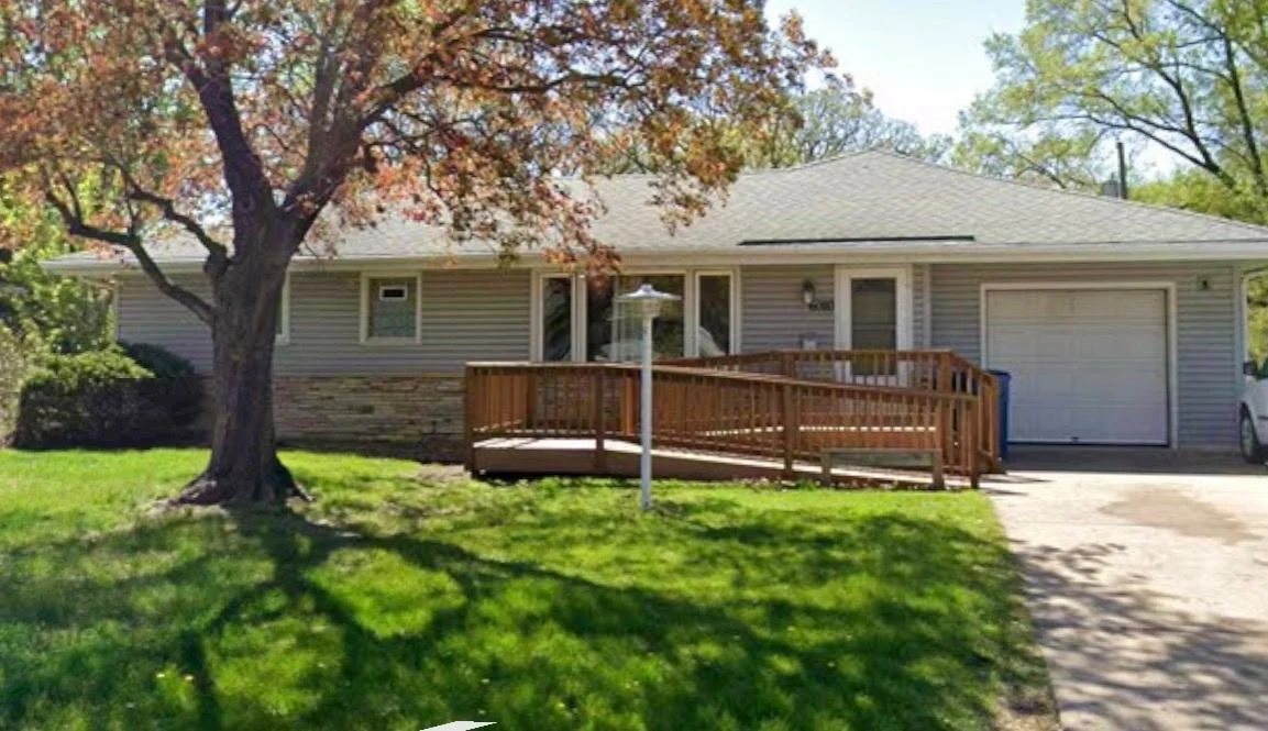 Grey single-story house with a front yard, tree, and driveway. There is a wooden ramp leading to the front door, a lamp post in the yard, and a garage on the right side.