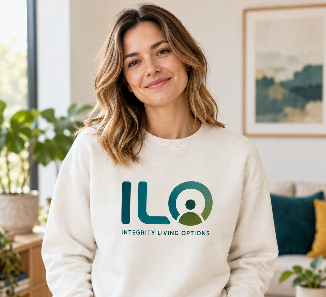 Smiling woman wearing a white sweatshirt with ILO logo and 'Integrity Living Options' text, standing in a bright living room with plants and artwork.