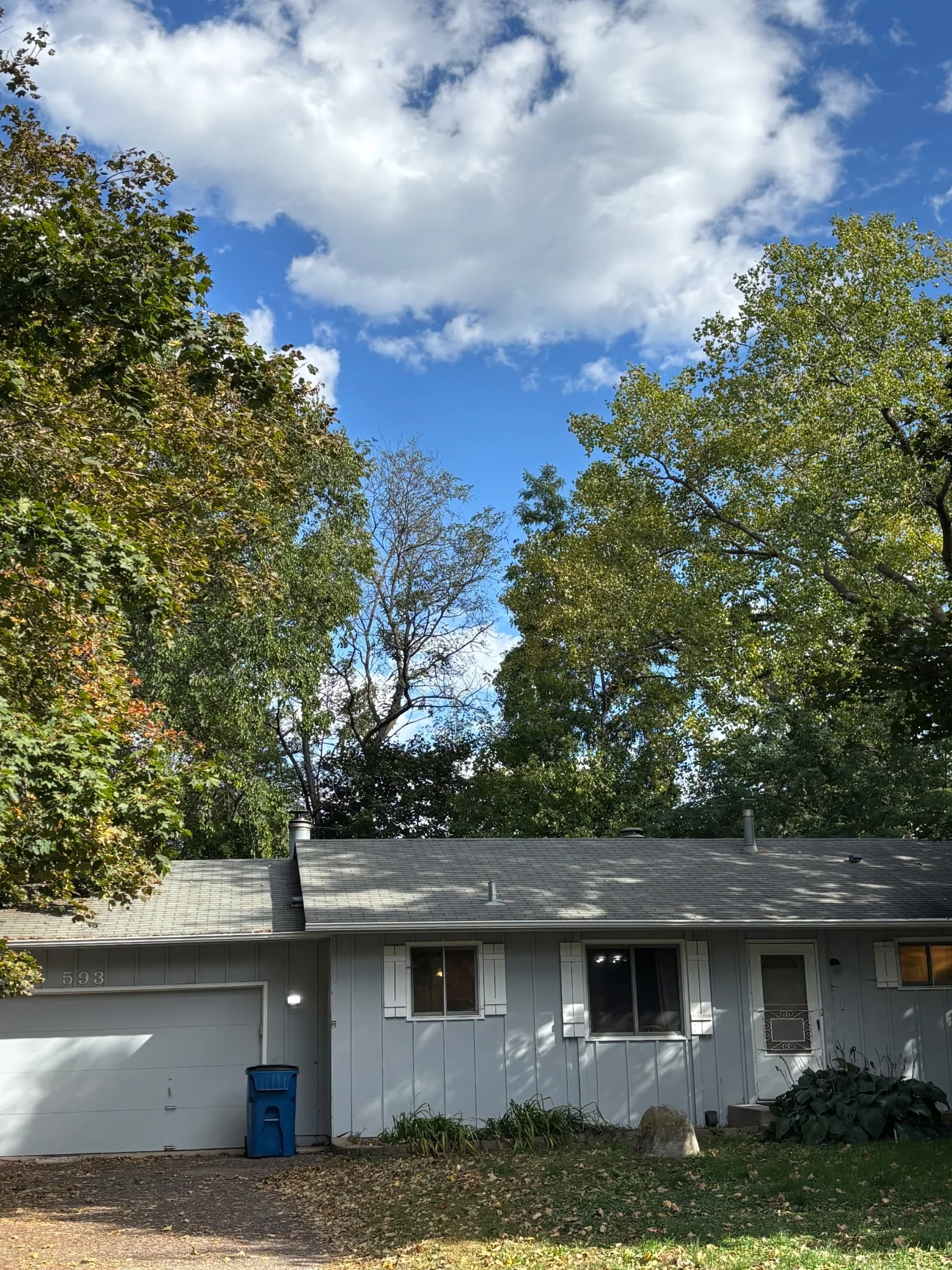 A house with a gray roof and white siding, surrounded by trees with green and some yellowing leaves, under a blue sky with scattered white clouds.