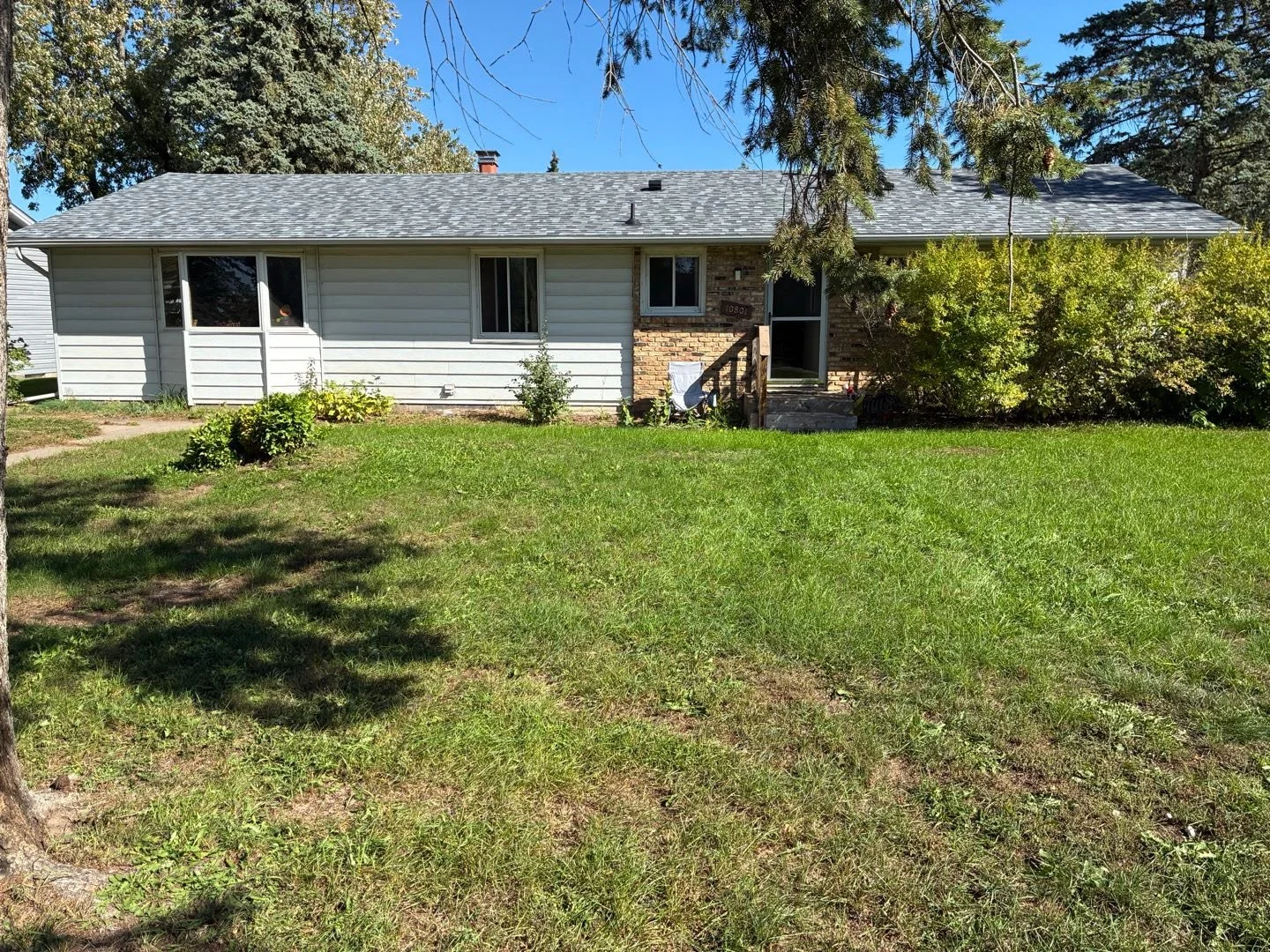 A single-story house with white siding and brick exterior, surrounded by a green lawn and bushes. The house has a small porch with steps leading to a front door, and several windows. Trees are visible in the background under a clear blue sky.