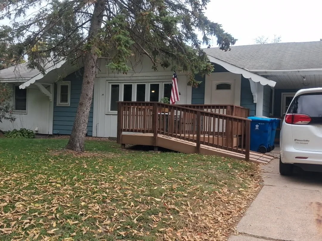 A white house with a wooden ramp leading to the front door, an American flag hanging near the entrance and a white vehicle parked in the driveway. There are fallen leaves on the yard and a large tree in front of the house.