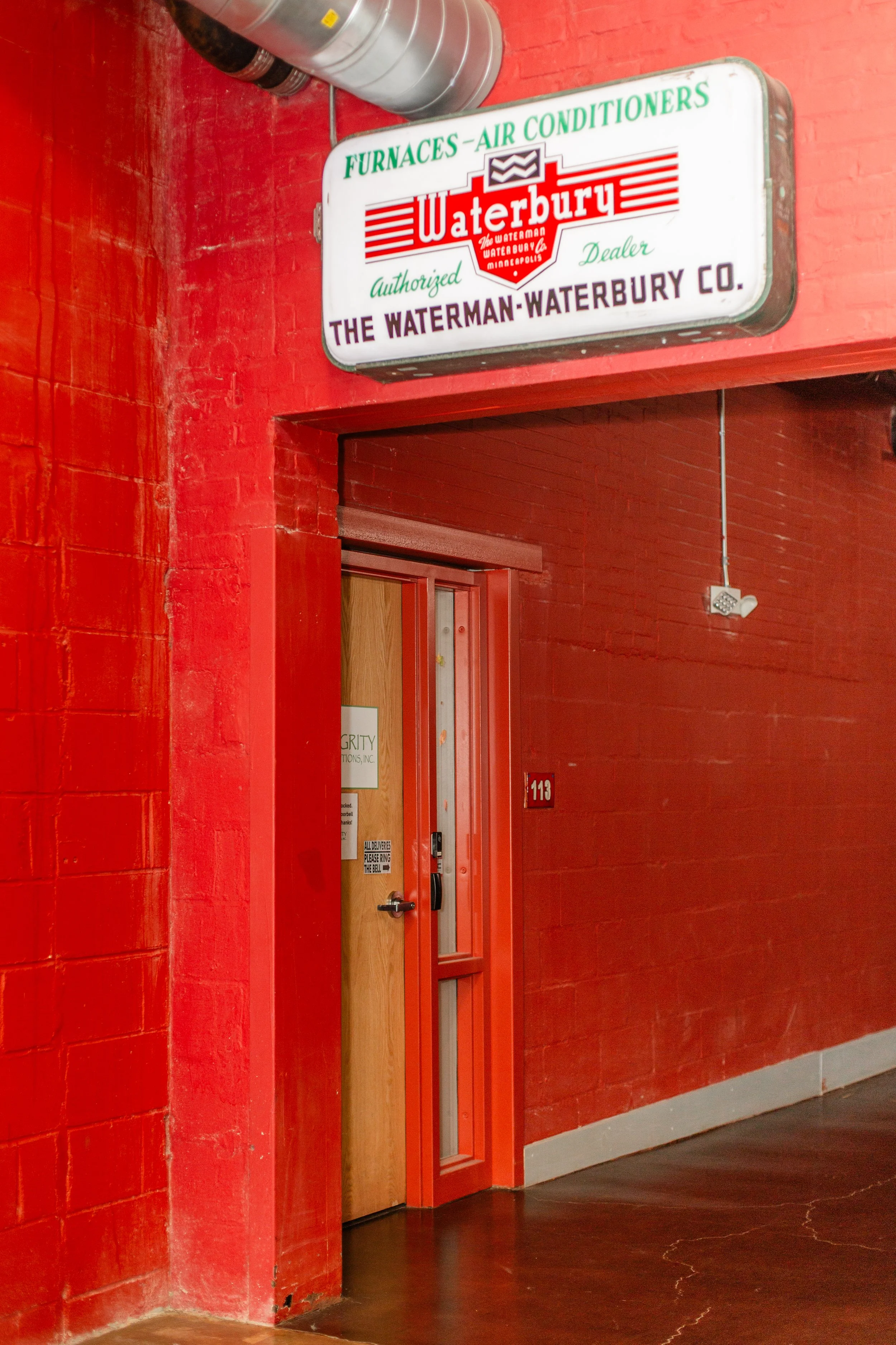 A red brick hallways with a mounted Signboard with green, red, purple, and white text displaying the name Waterbury. In the hallway is a wooden door with signs for Integrity Living Options. 