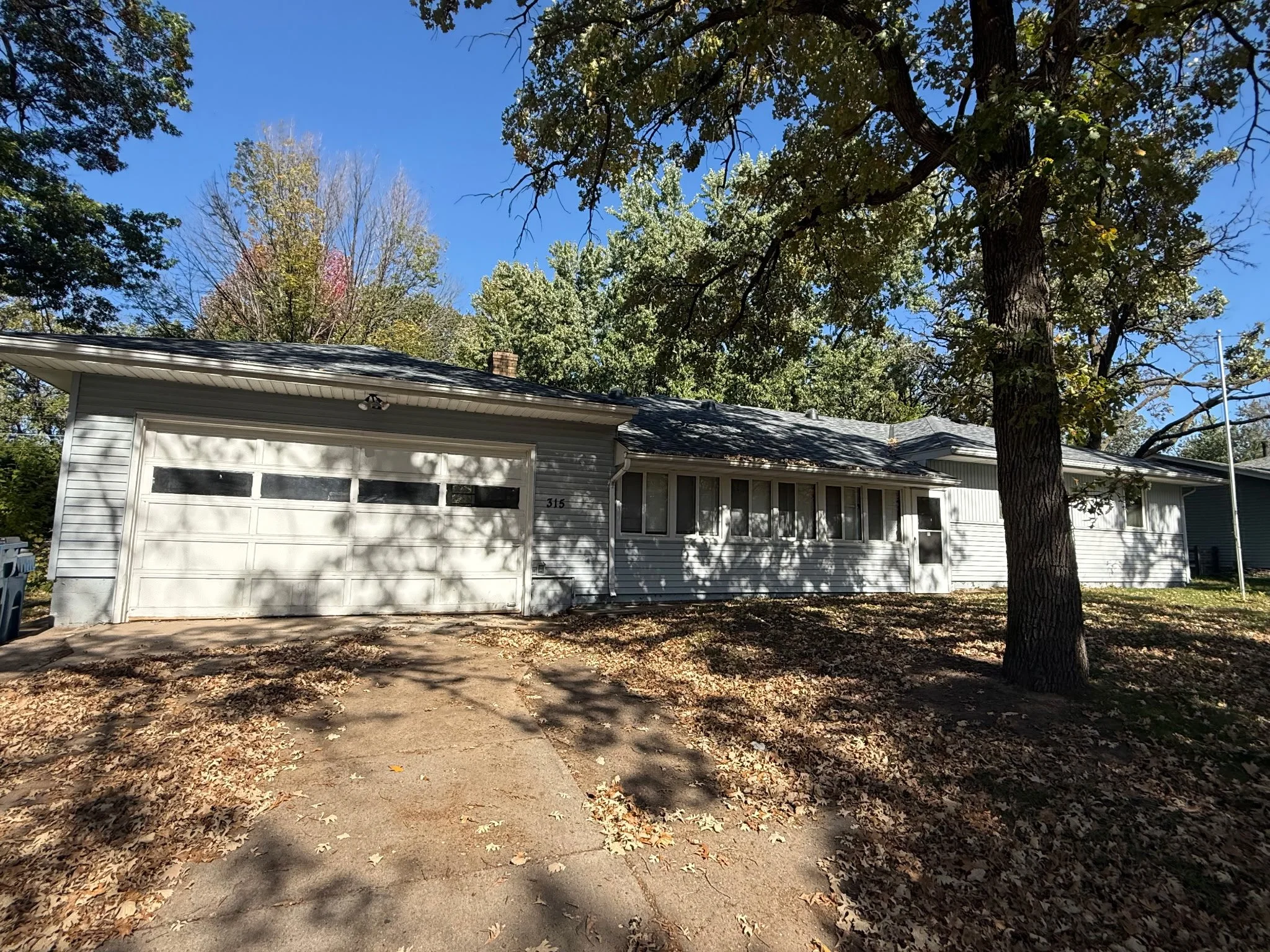 A suburban house with a white exterior, garage door, and front porch, surrounded by trees with green and yellow leaves, under a clear blue sky, with fallen leaves on the ground.