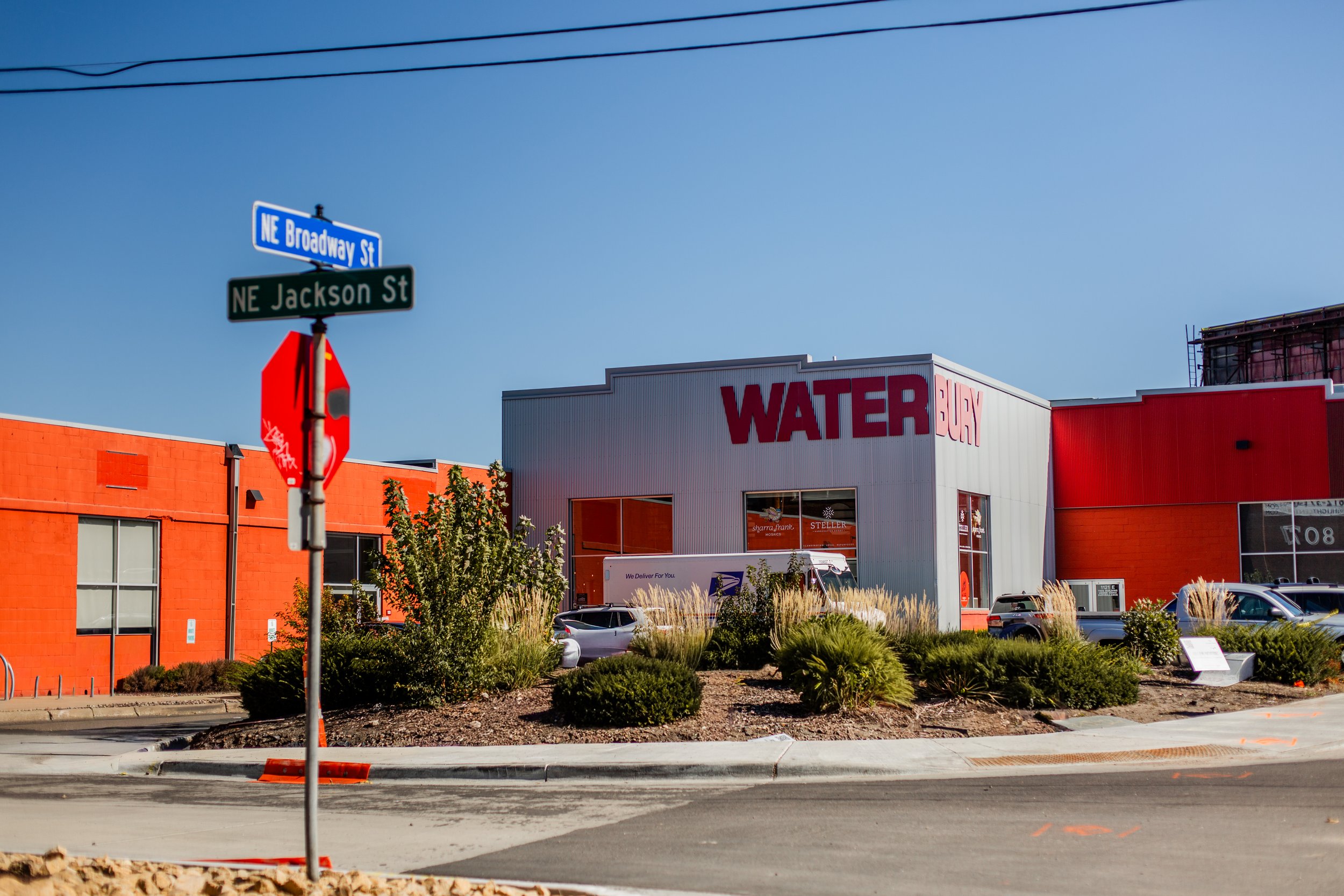 Street view showing traffic signpost with street names NE Broadway St and NE Jackson St, a building with large red sign that says "WaterBury", parked cars, landscaping with bushes and trees, and a clear blue sky.
