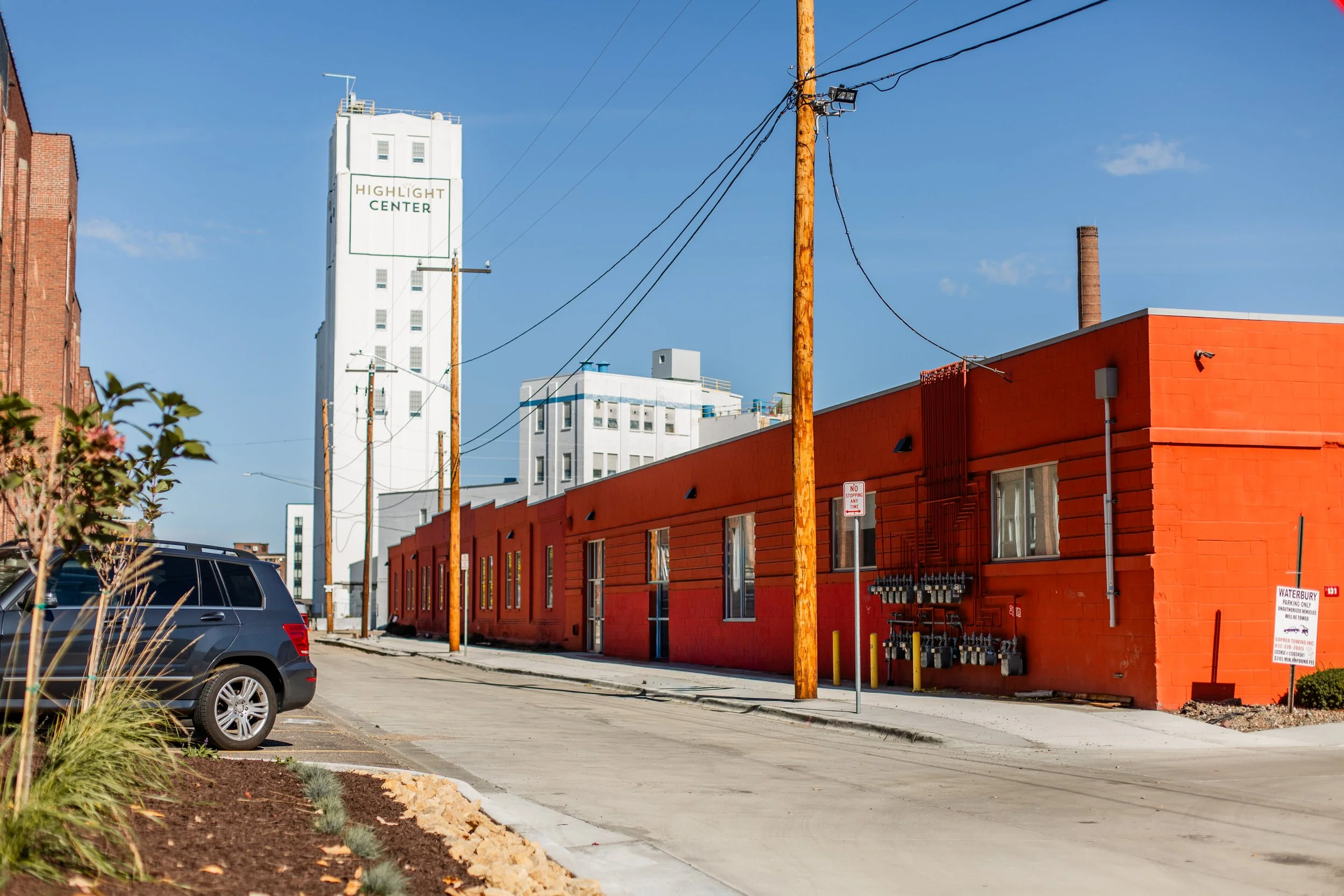 A street scene with a red building on the right, utility poles and wires, a parking lot with a black SUV, and white buildings in the background, under a clear blue sky.