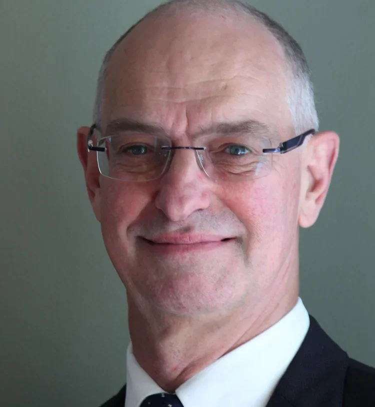 Close-up portrait of a middle-aged man with glasses, short gray hair, and a friendly smile, wearing a suit and tie against a plain background.
