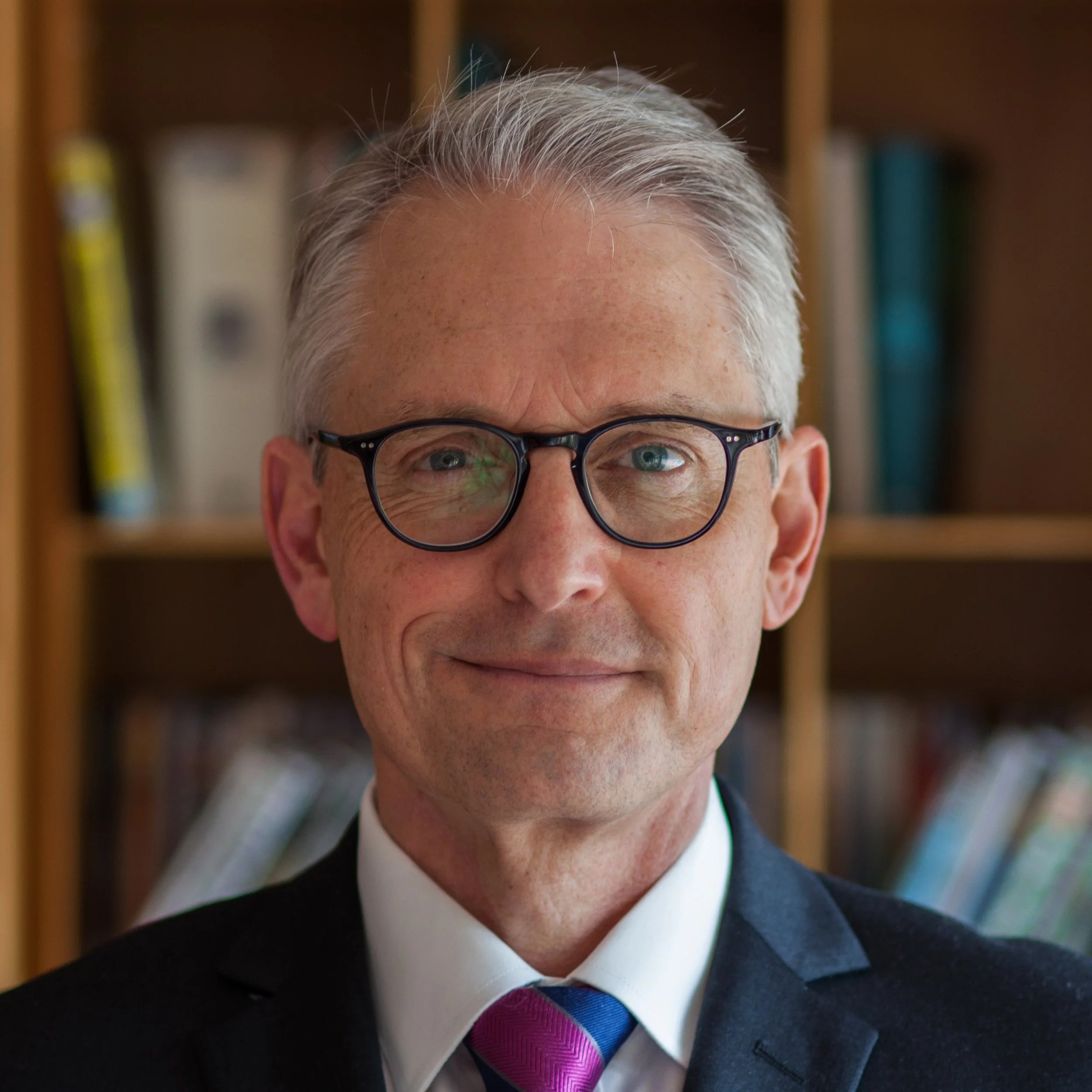 Close-up of a man with gray hair, glasses, smiling, dressed in a dark suit, white shirt, and a pink and blue striped tie.
