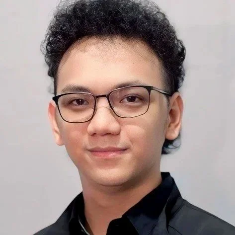 Headshot of a young man with curly dark hair, wearing glasses, and a black collared shirt, smiling subtly against a plain background.
