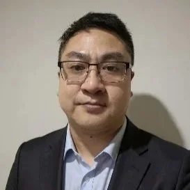 A man wearing glasses, a dark suit, and a light blue shirt, standing against a plain background.
